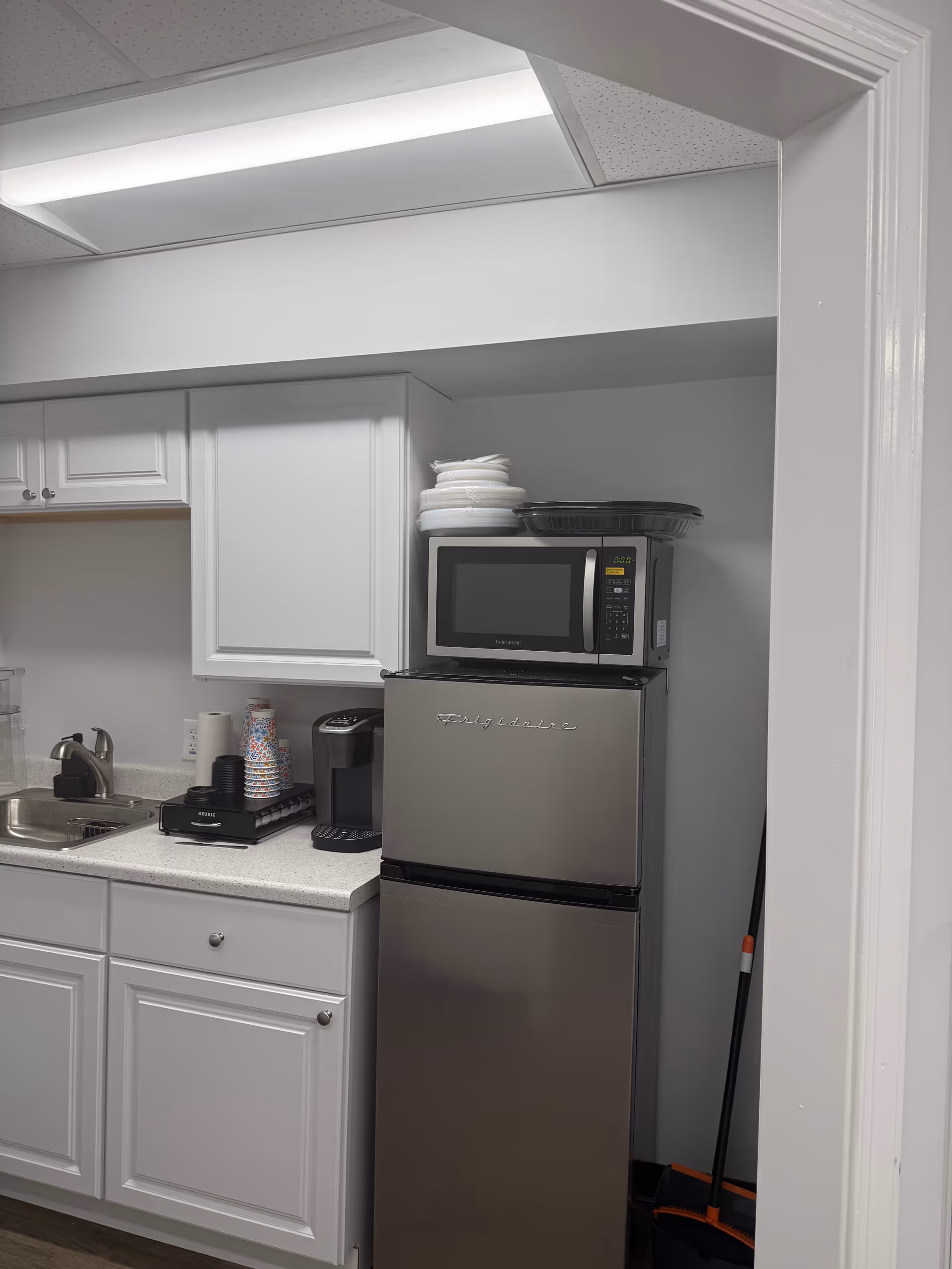 Compact kitchenette with white cabinets, microwave atop a stainless refrigerator and coffee maker.