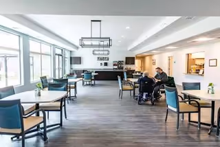 Bright interior dining room with tables, chairs, large windows, and a few residents seated.