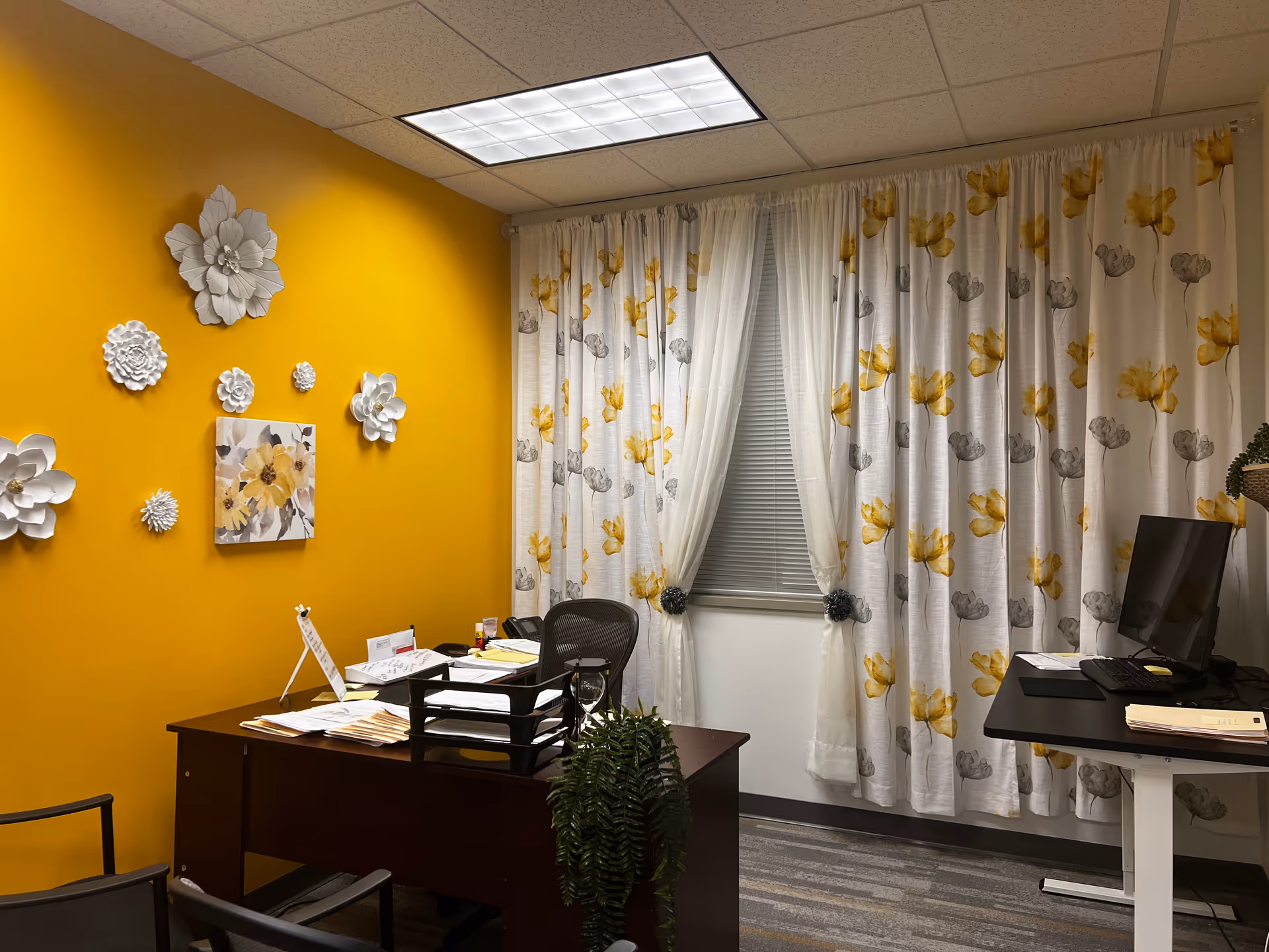 Office workspace with yellow accent wall, framed flower decor, window with patterned curtains, and a desk.