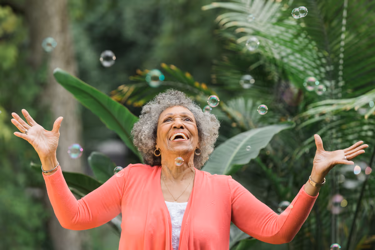 An elderly woman outdoors raises her arms with joy among bubbles and lush plants.