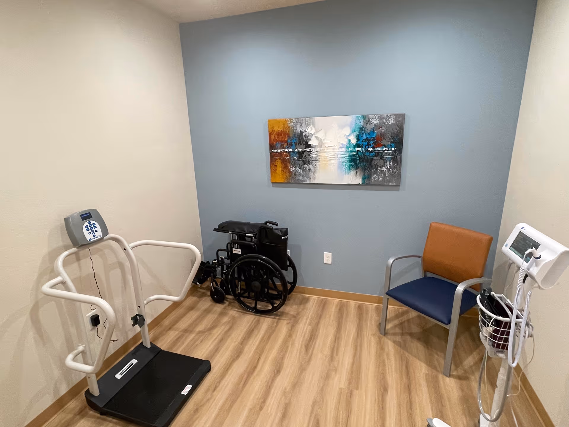 Small clinical room with a wheelchair, medical equipment, a chair, and abstract wall art.
