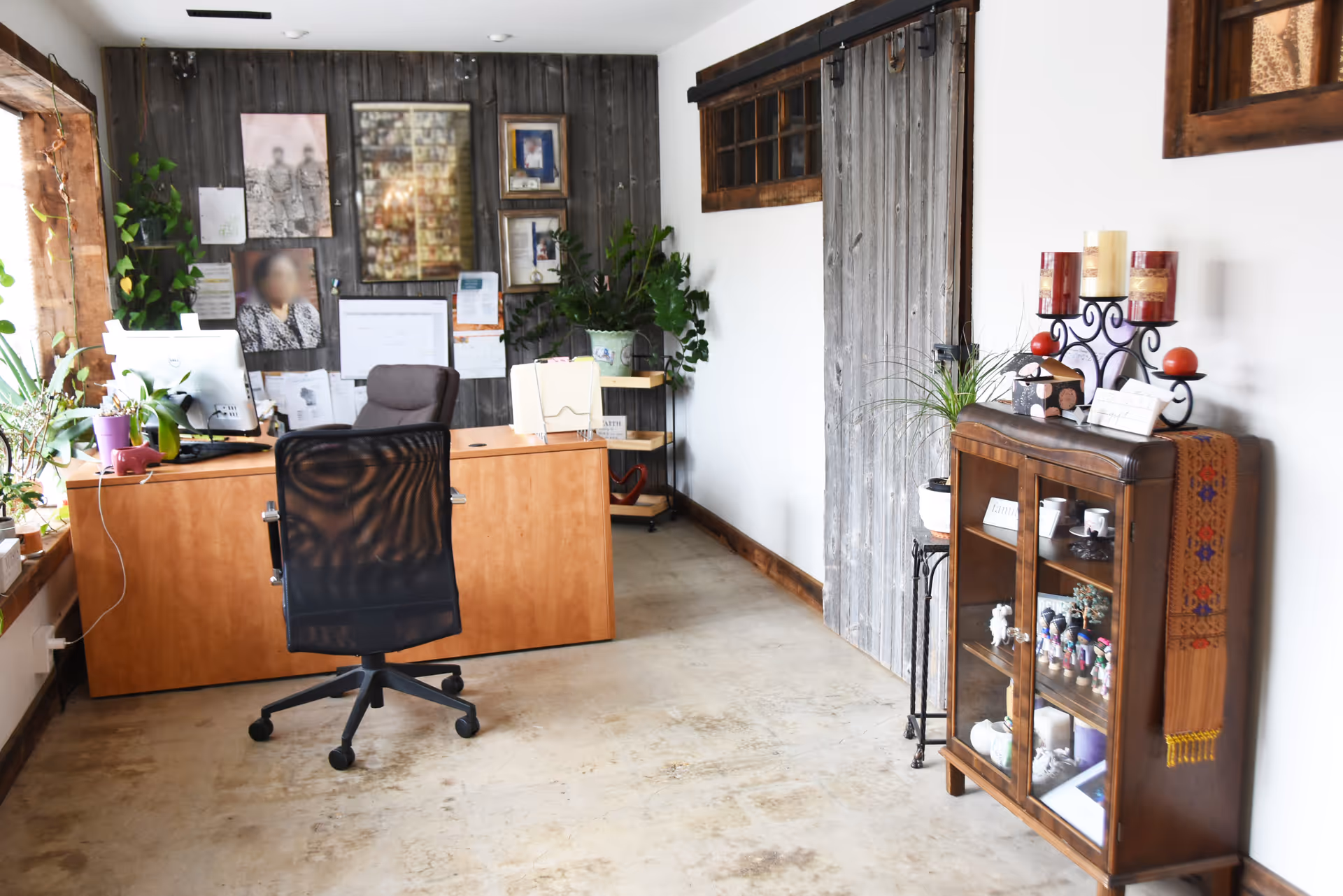 An office workspace with desk, swivel chair, framed photos, and wooden display cabinet.