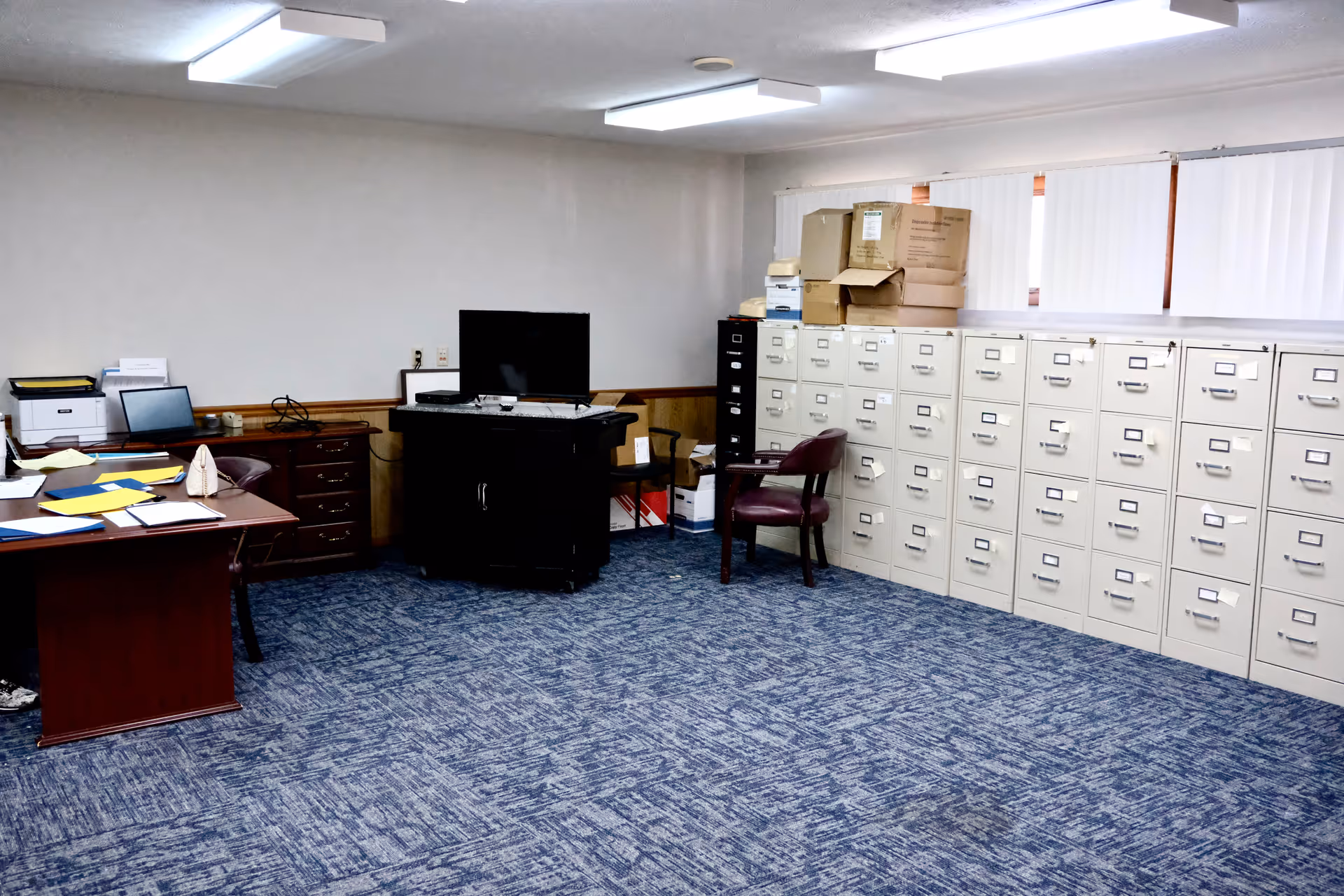 An office workspace with a desk, computer, filing cabinets along the wall, and stacked boxes.