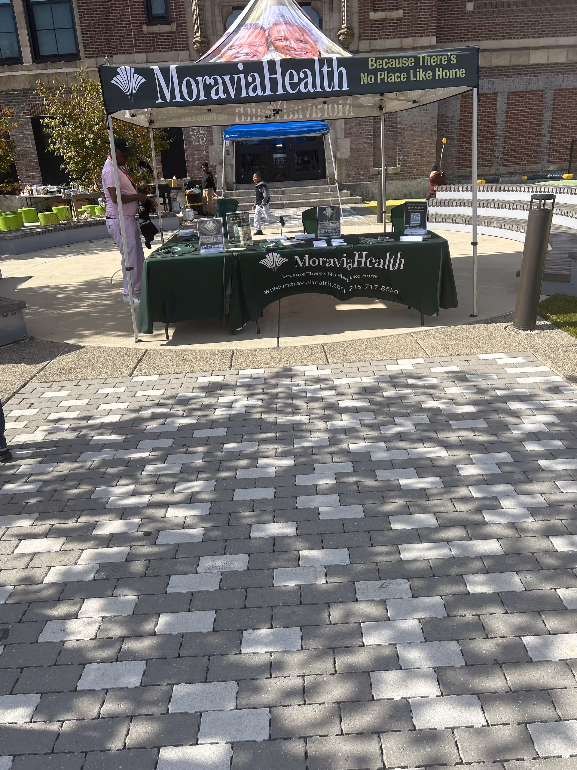 An outdoor Moravia Health booth with banners, a table, and people nearby.