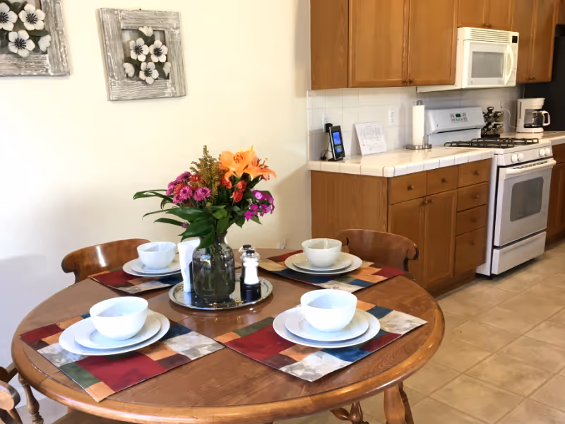 A round dining table with place settings and a flower centerpiece in a kitchen dining area.