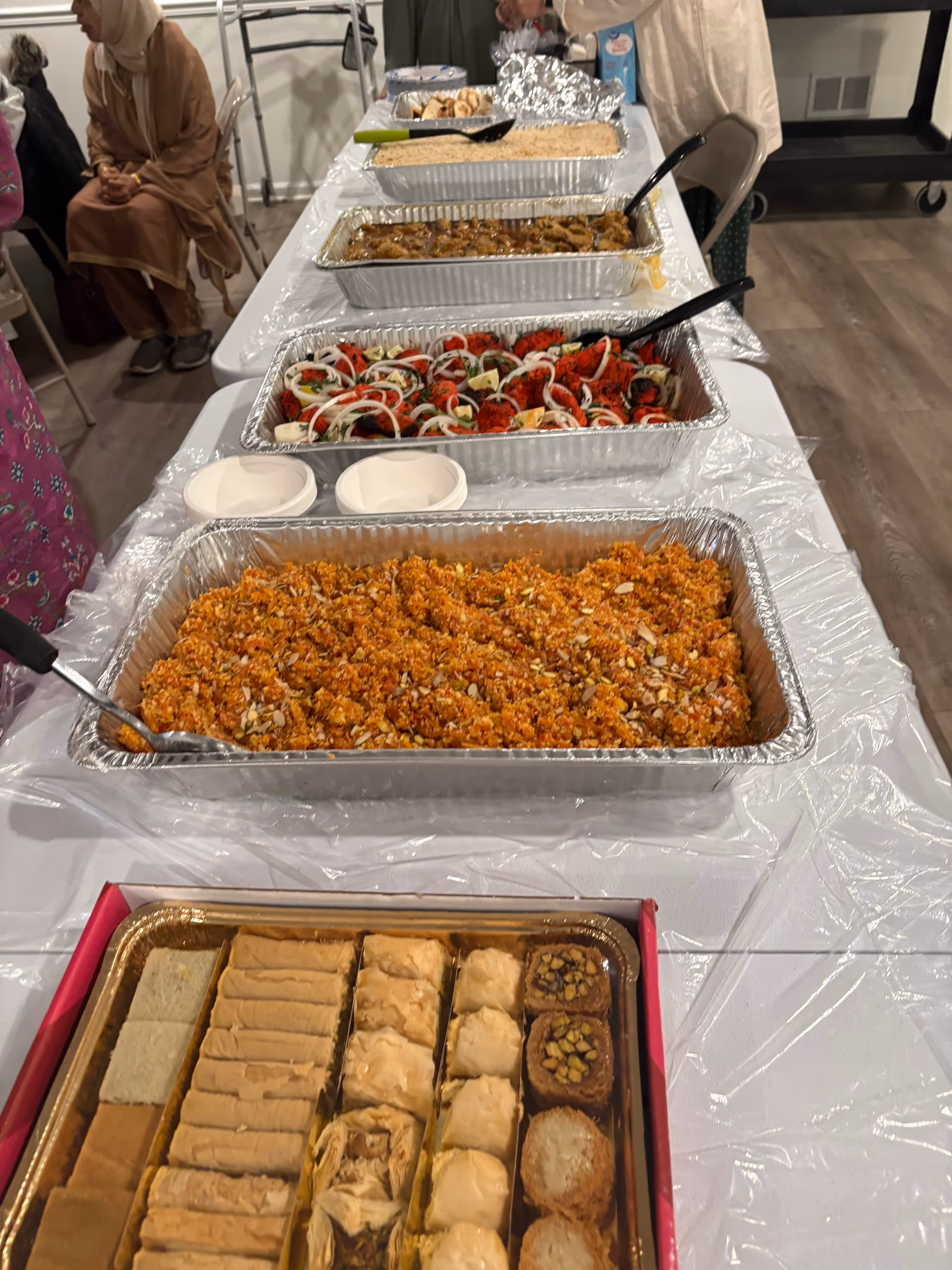 Buffet-style food trays line a long table in a senior care dining area, with guests nearby