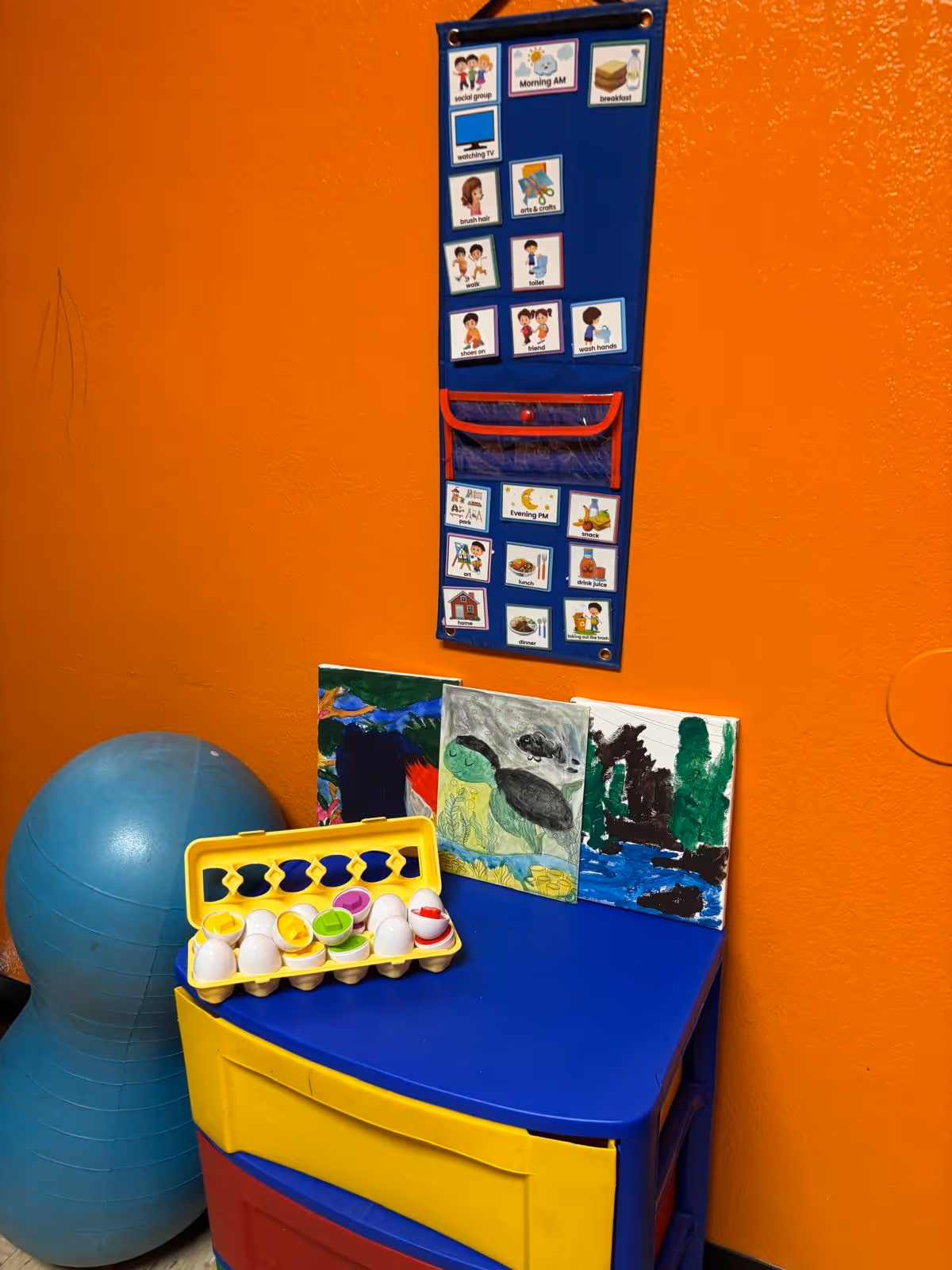 Bright orange wall with a blue activity chart, artwork, and colorful storage in an interior corner.
