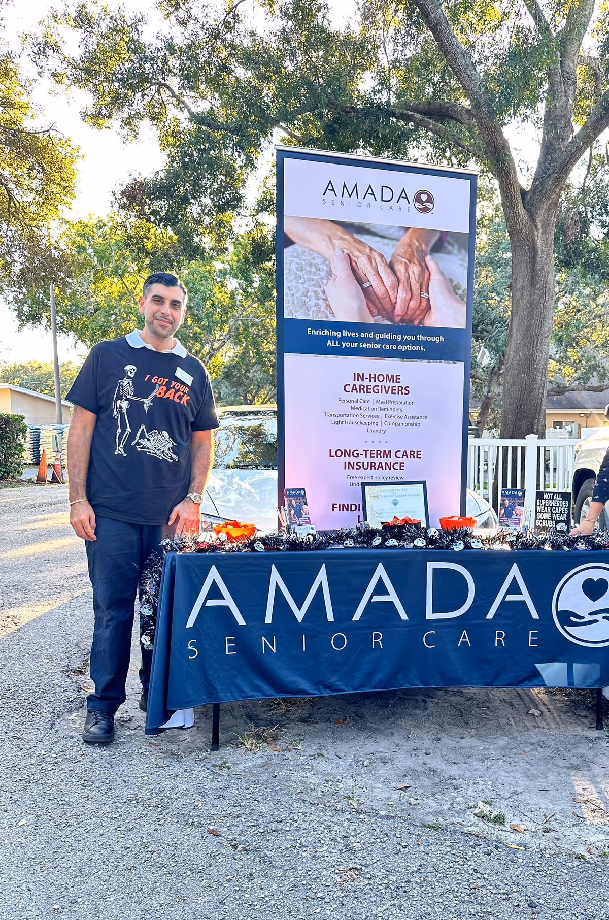 A man stands beside an Amada Senior Care banner at an outdoor event.