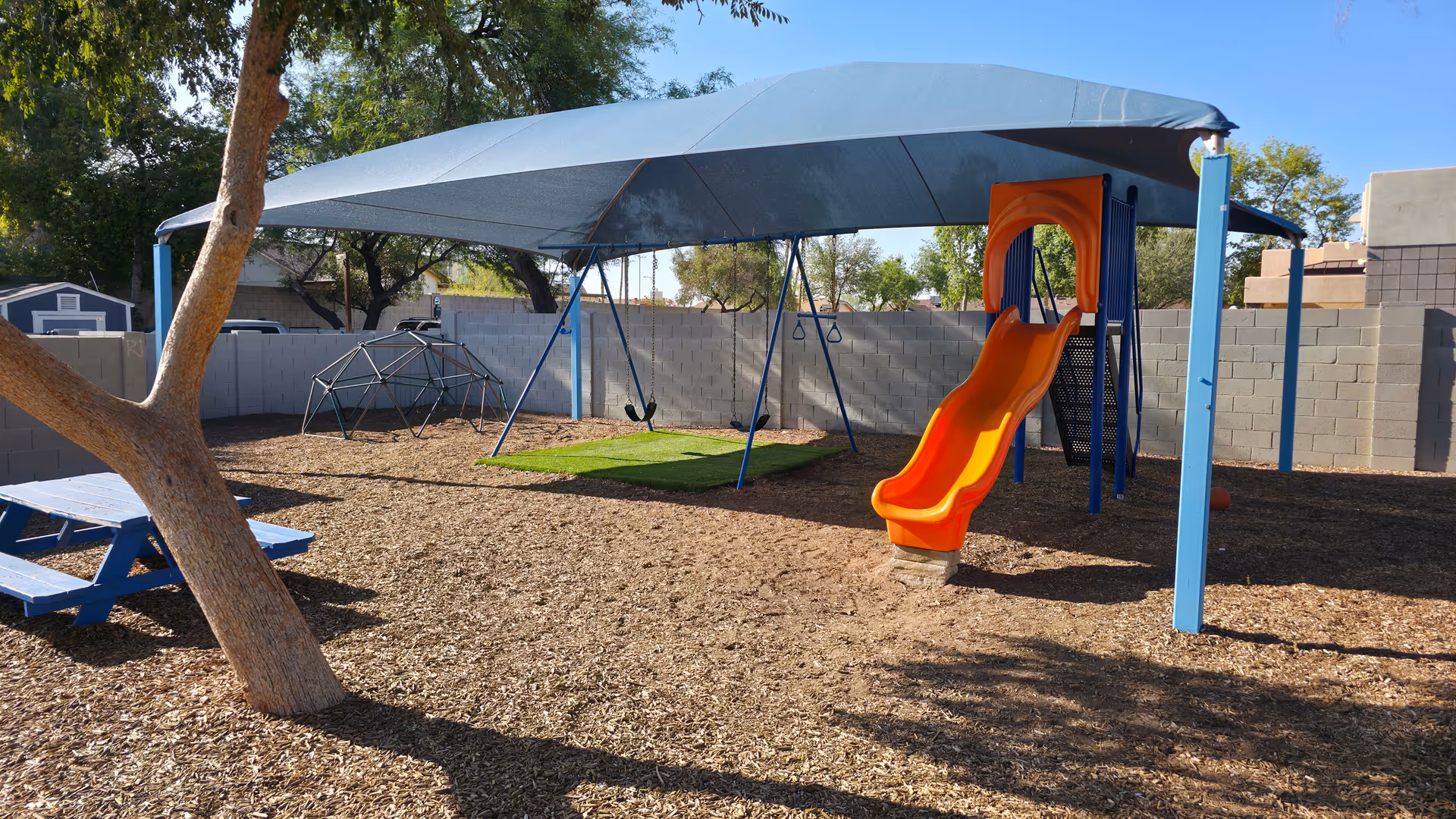 Outdoor playground area with a shade canopy, swings, and a bright orange slide.