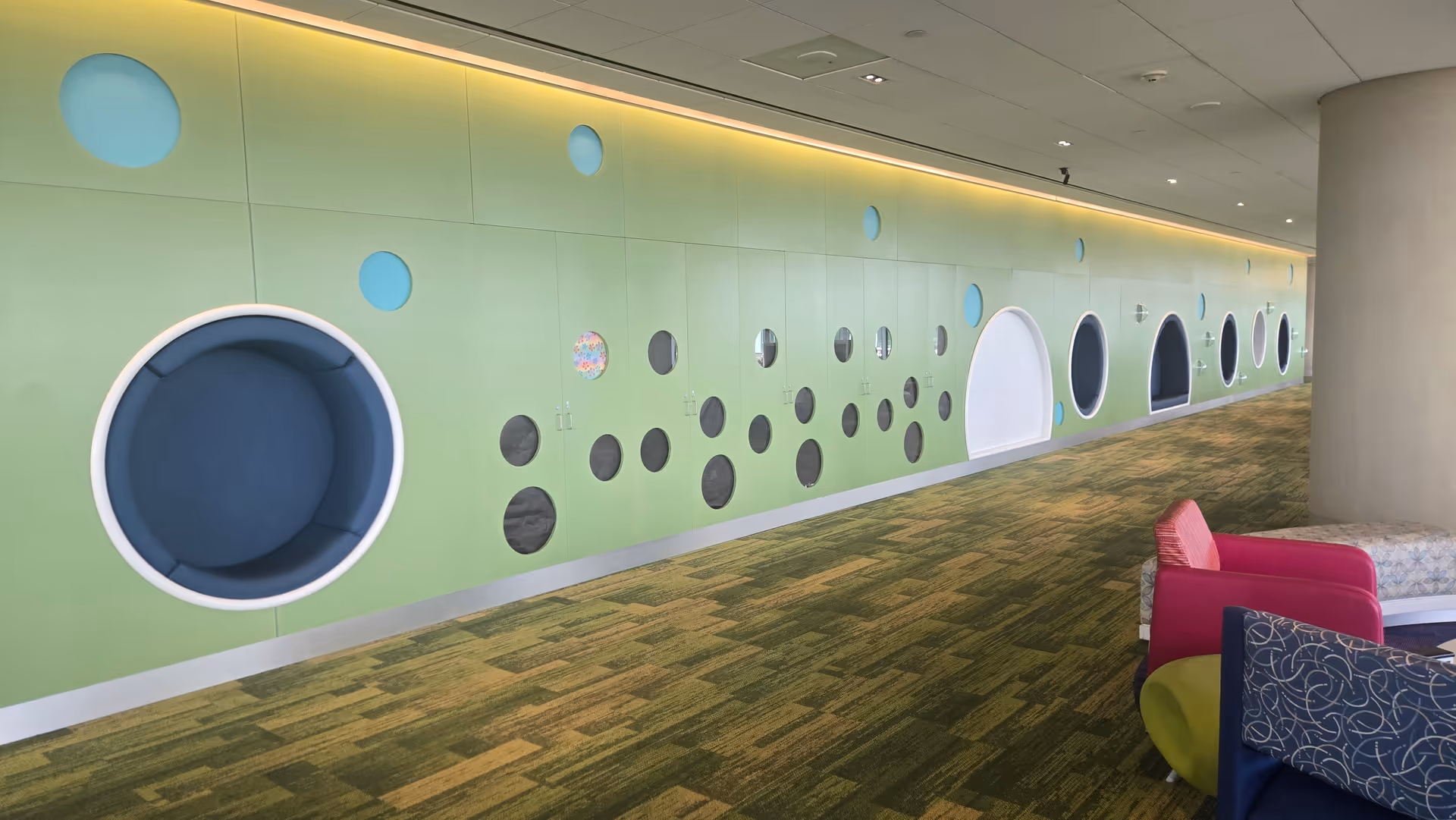 Long interior hallway with lime-green wall and circular wall seats; colorful lounge chairs on right.