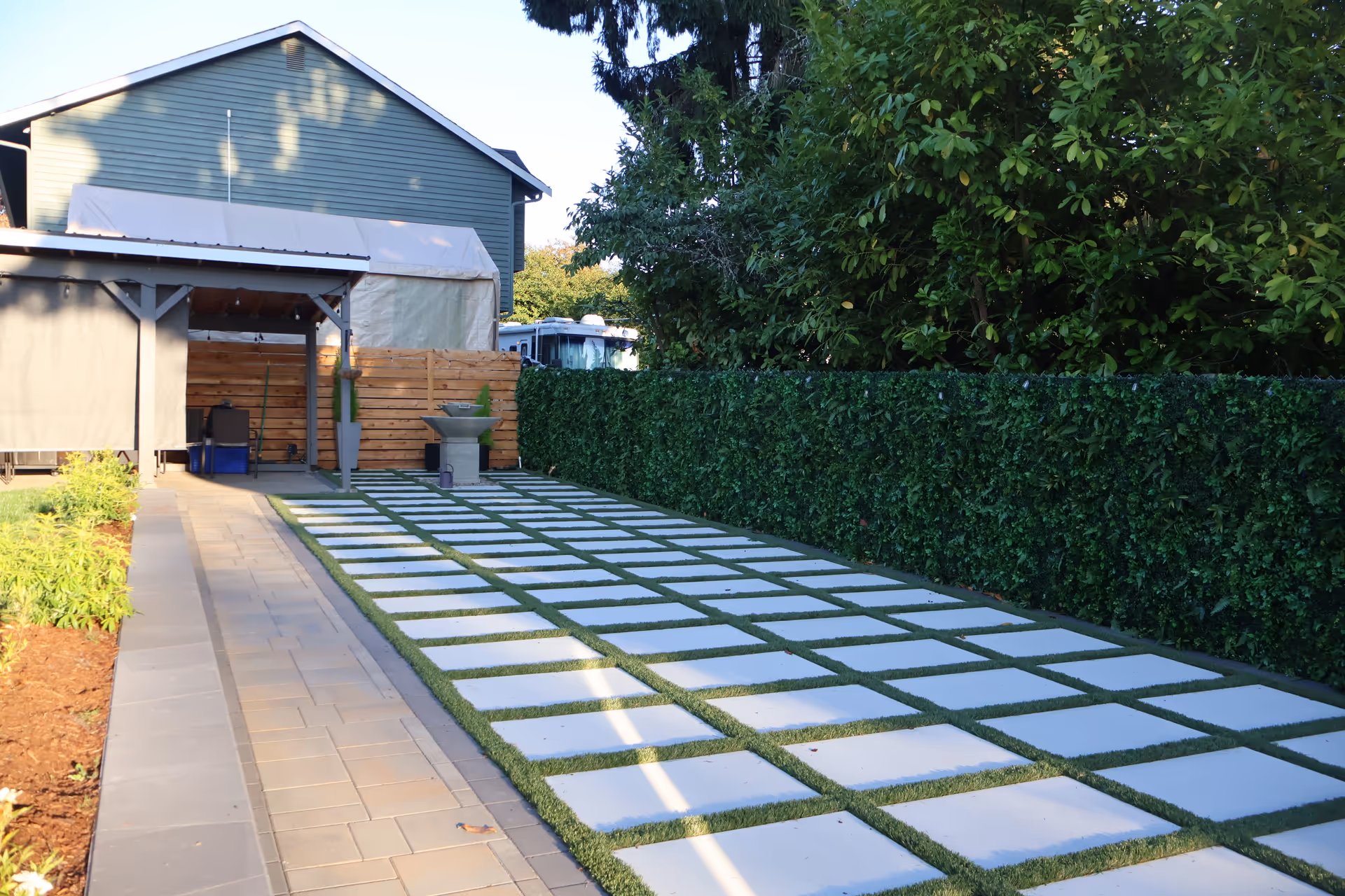 Outdoor pathway with square stepping stones, hedge, and a covered seating area.