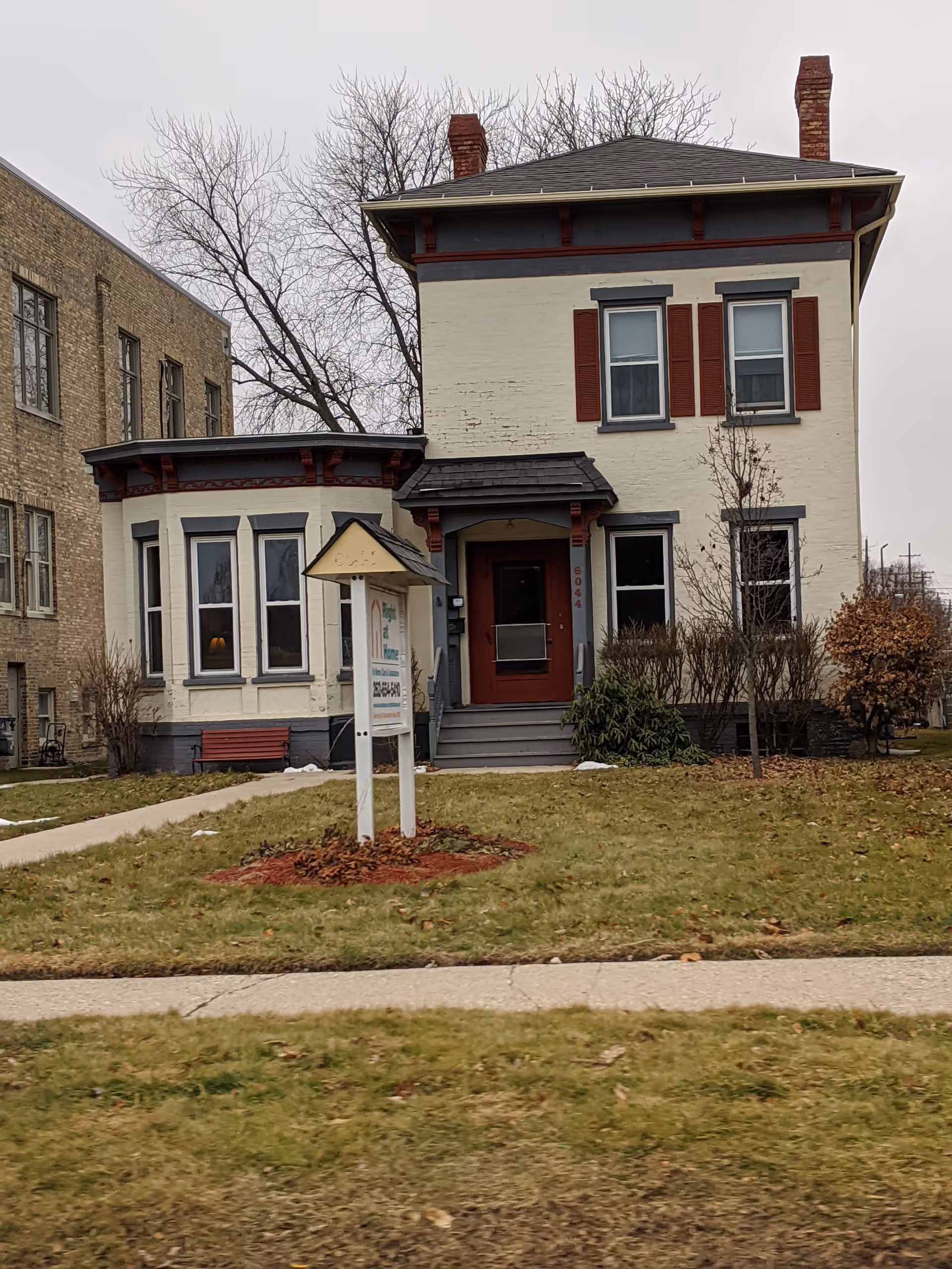 A two-story beige building with red-trimmed windows, front porch, and a sign near the lawn.