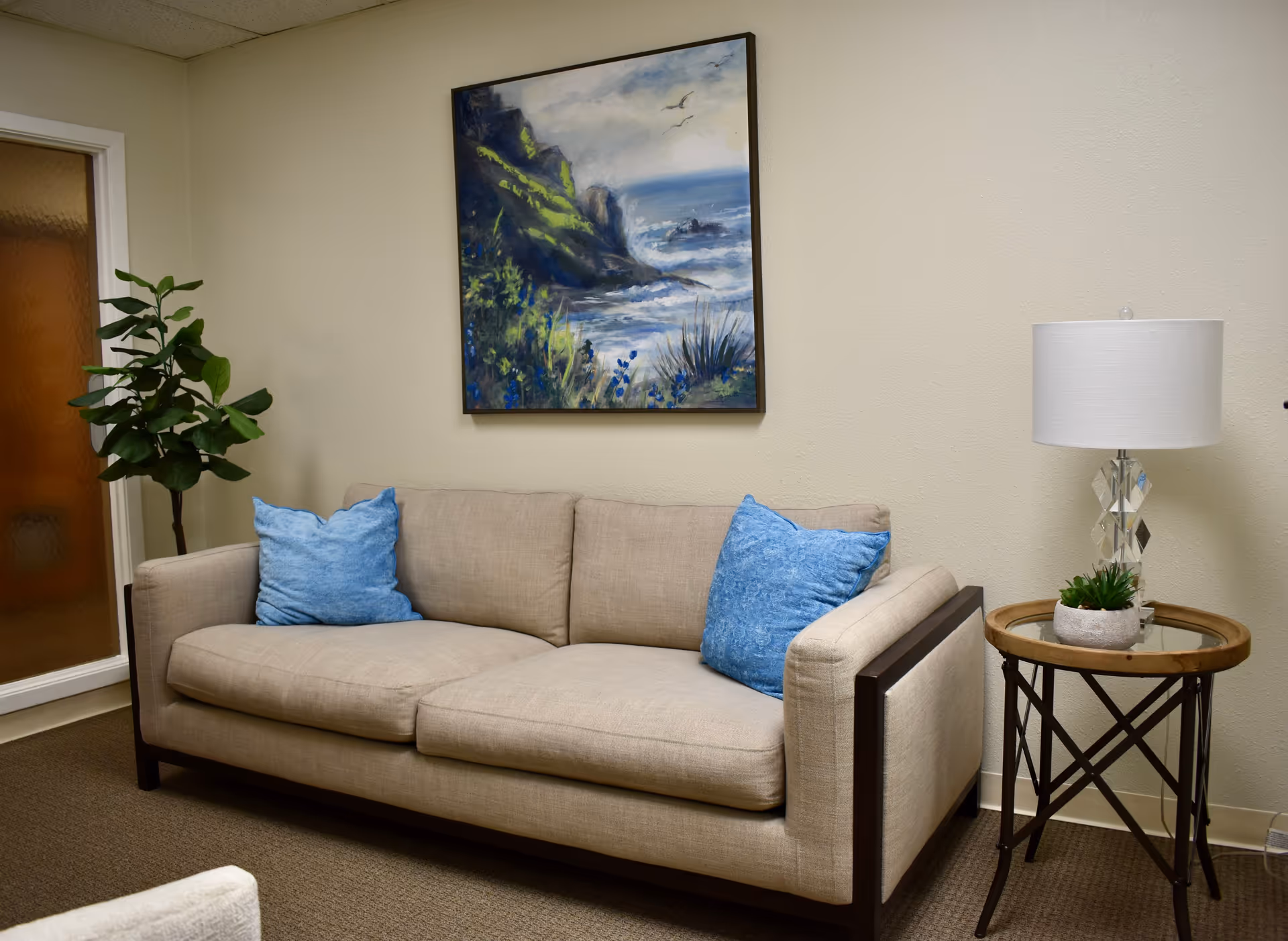 Beige living room with sofa, blue cushions, wall art, plant, and side table lamp.