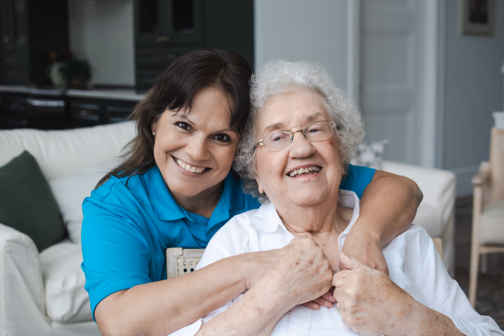A caregiver hugs a smiling elderly resident in a bright living room.