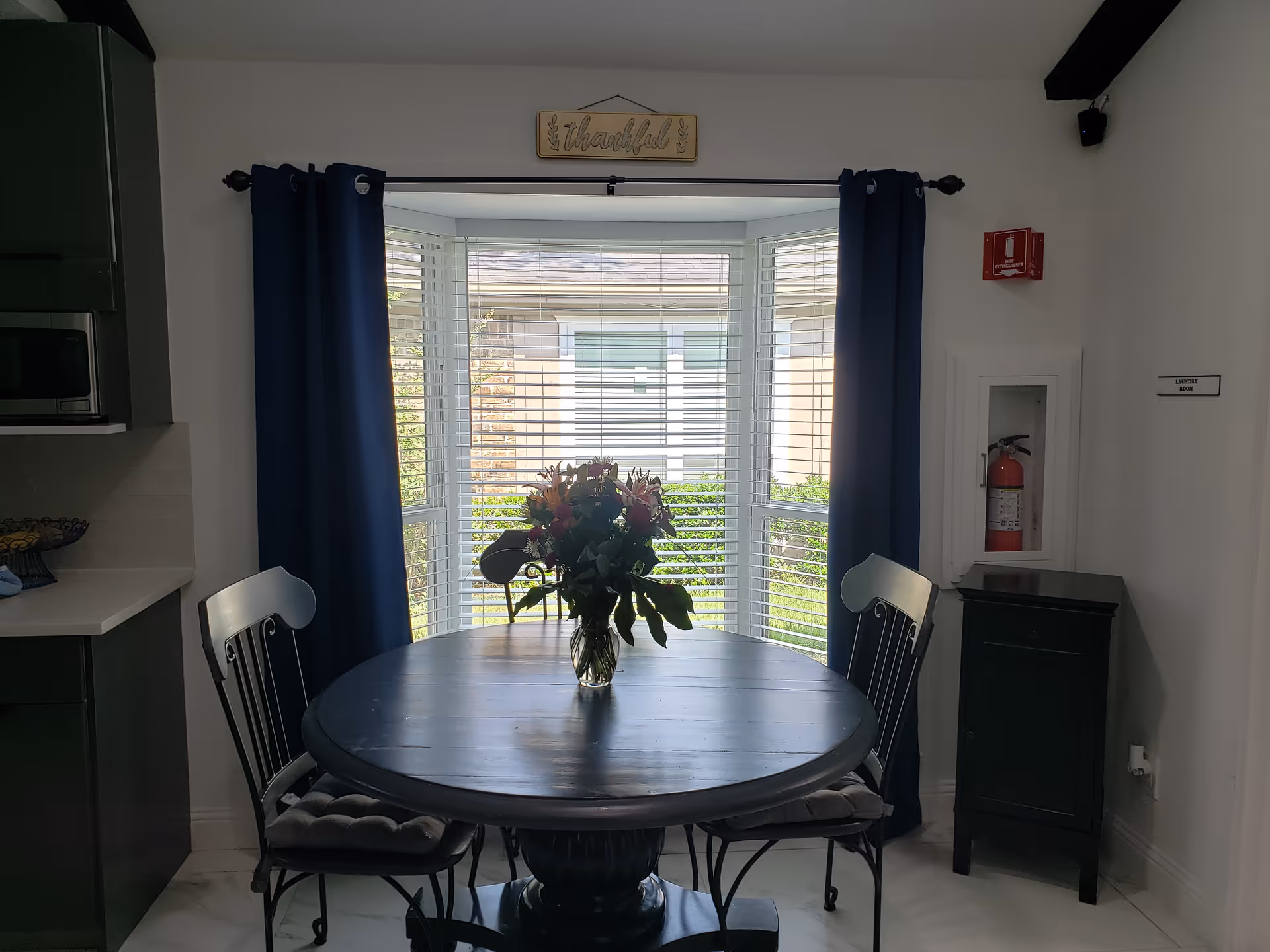Dining area with round table, blue curtains, vase of flowers, and a kitchen to the left.