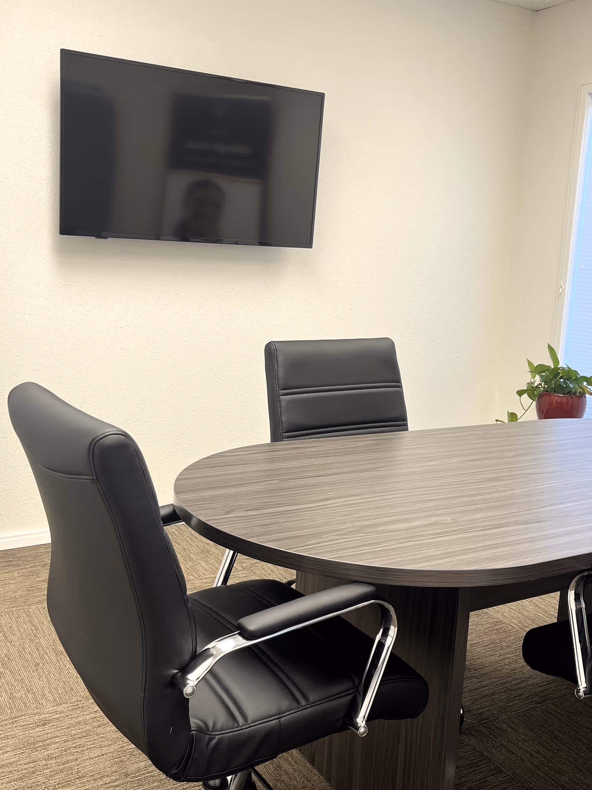 Modern conference room with a round table, black leather chairs, wall TV, and a plant.