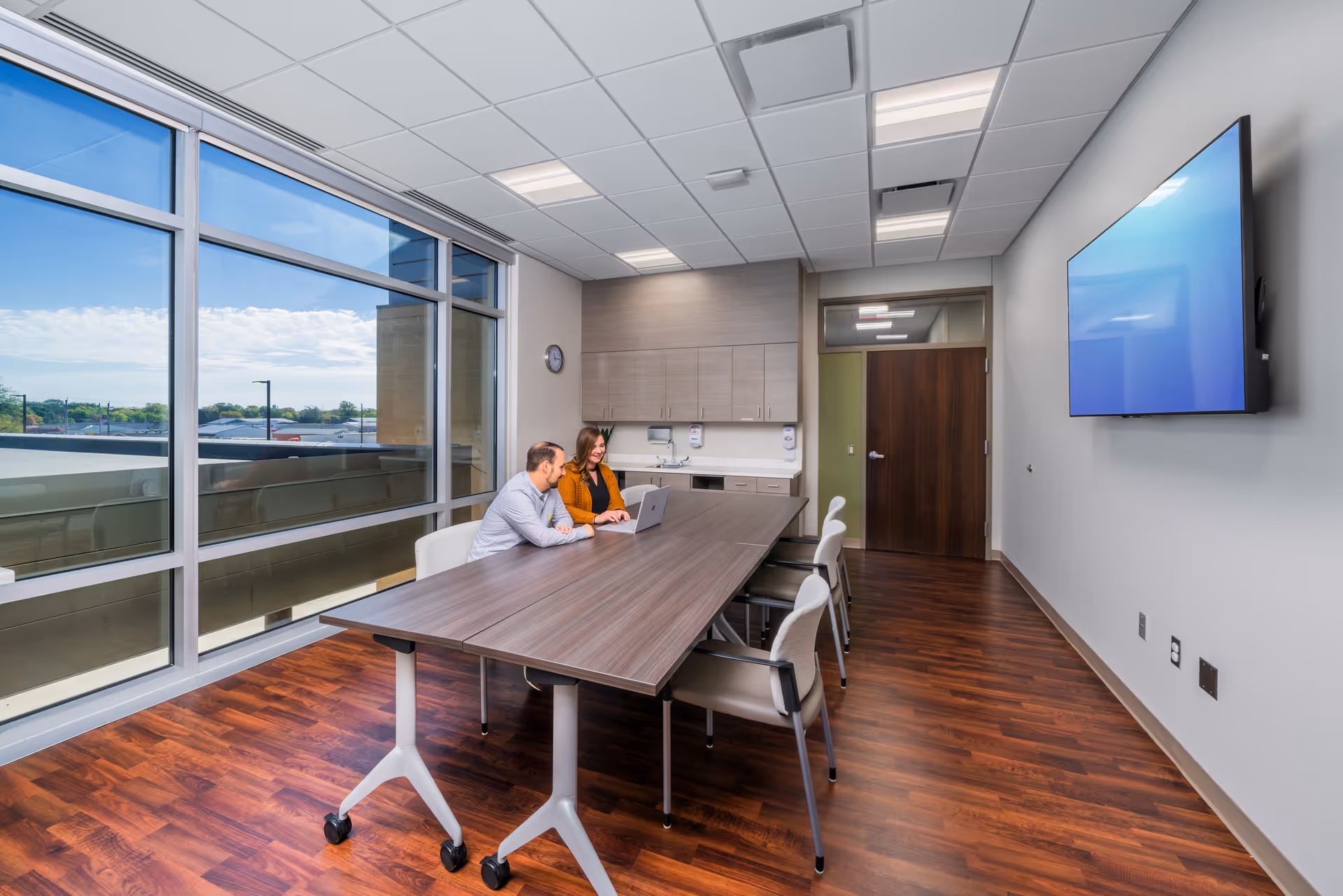 Two people sit at a long conference table in a modern meeting room with windows.