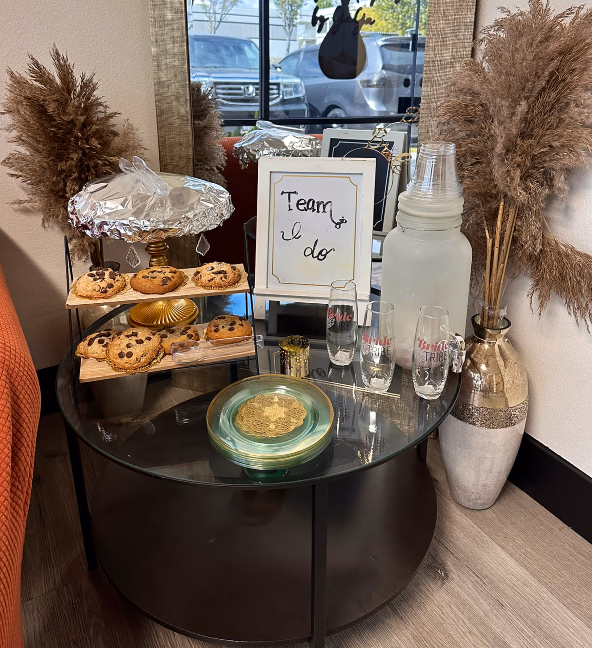 Round glass table with cookies, drinks, sign, and decorative plants in a cozy lounge.