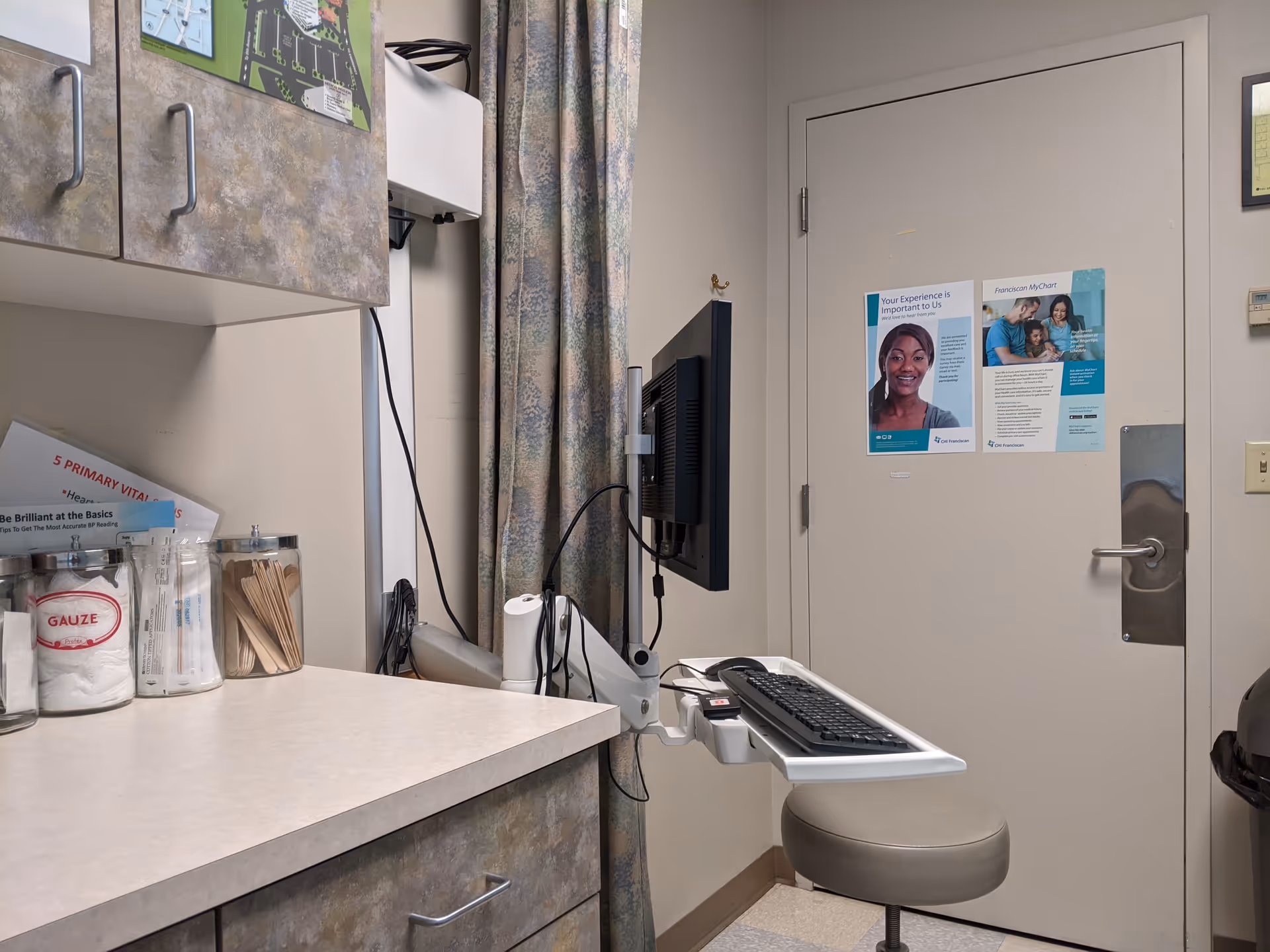 Clinic examination area with computer station, cabinets, a curtain, and posters on the door.