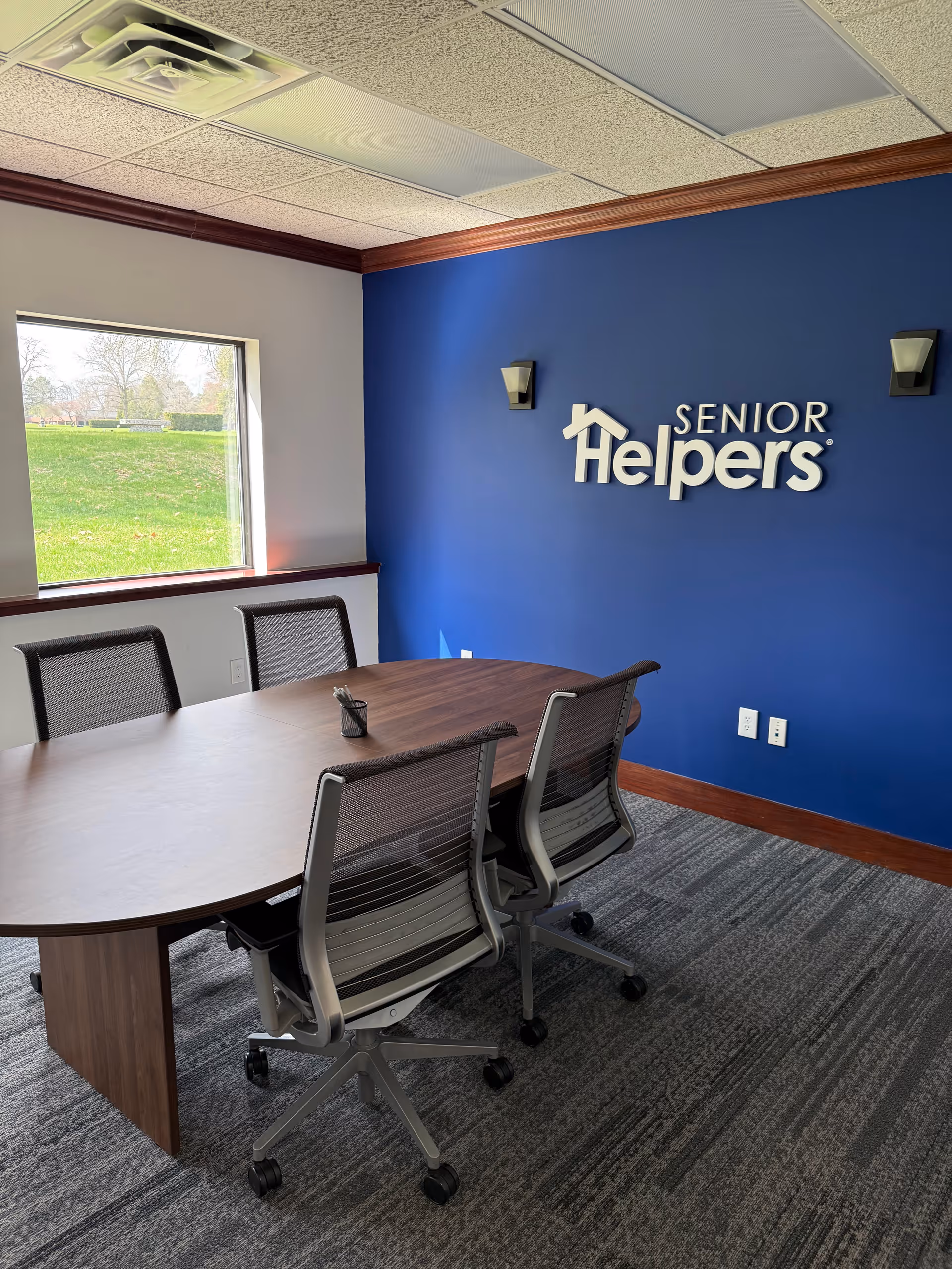 Meeting room with curved wooden table, office chairs, and a blue wall displaying Senior Helpers.