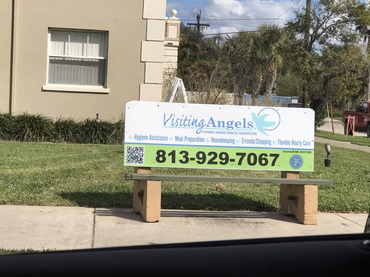 A large Visiting Angels sign on a bench outside a beige building with landscaping.