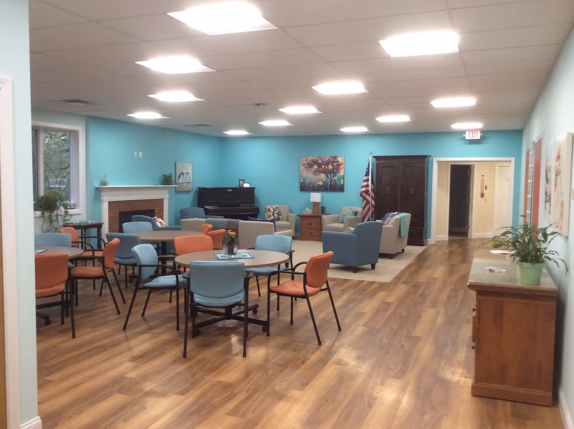 Bright teal-walled senior living lounge with multiple tables, chairs, and a piano.