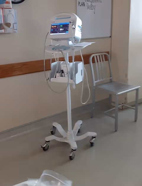Medical monitor on a wheeled stand in a hospital hallway beside a chair.