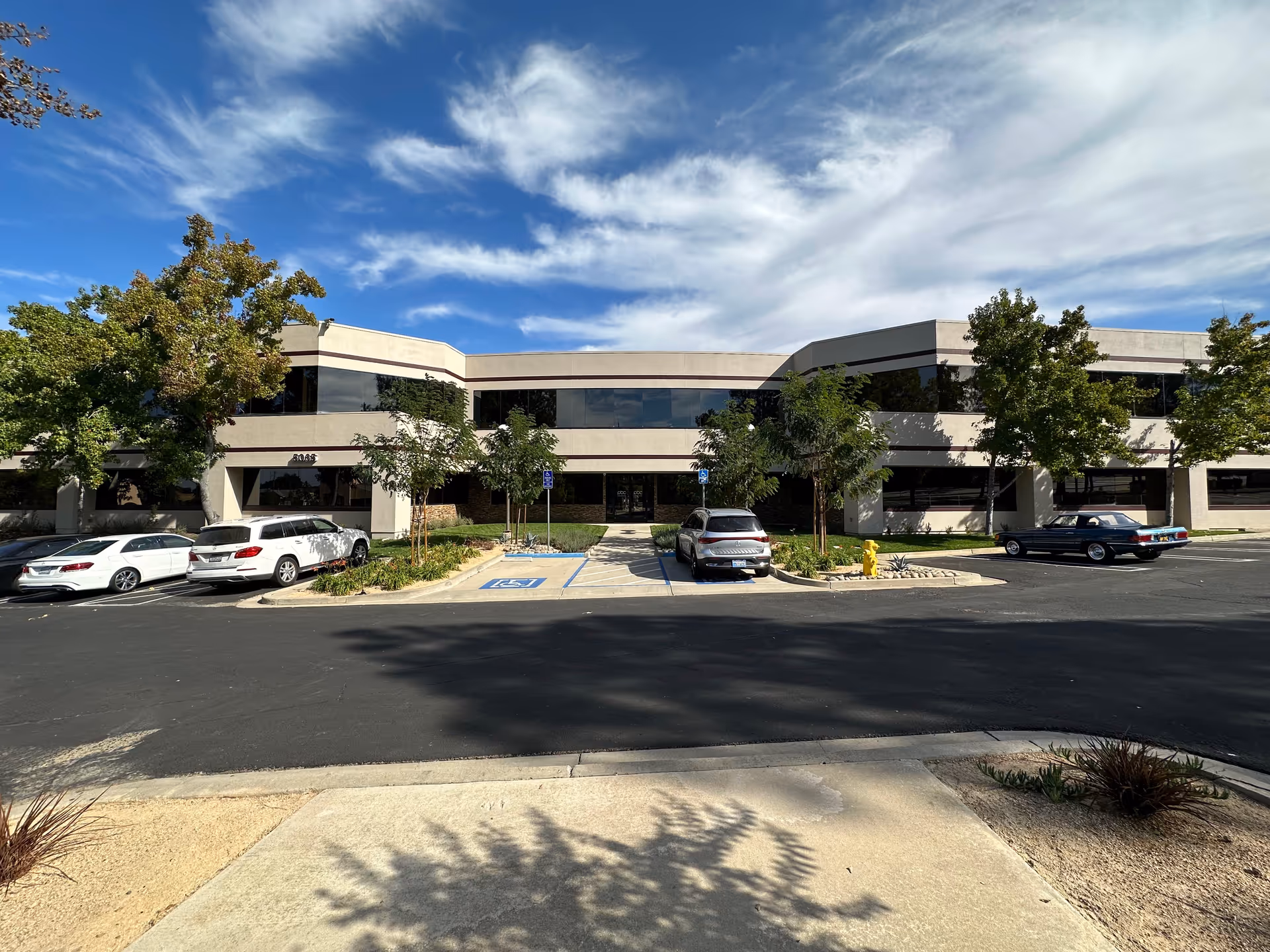 Two-story beige building with a parking lot and trees beneath a blue sky.