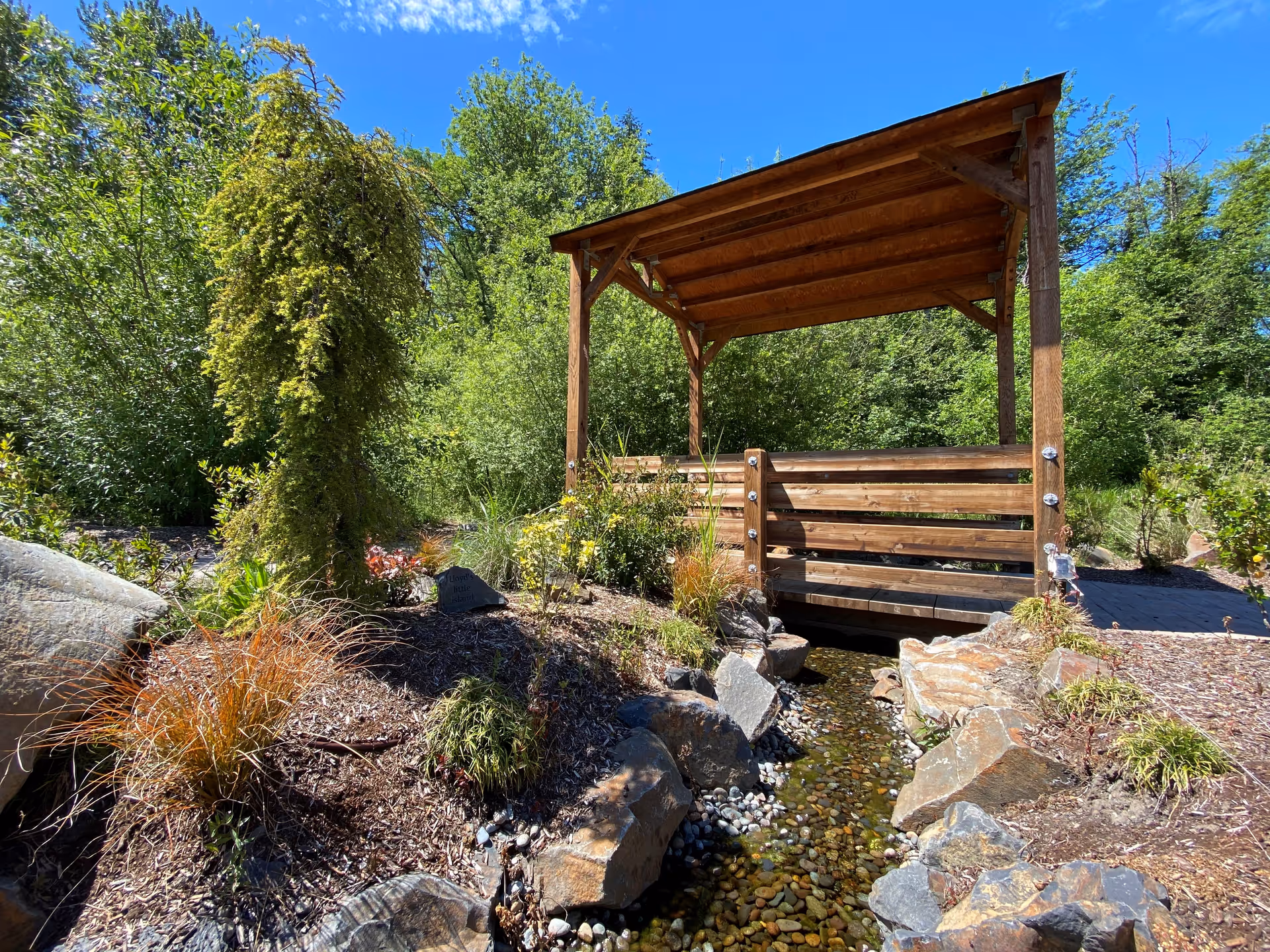 A wooden covered structure over a small stream surrounded by rocks and lush garden plants.