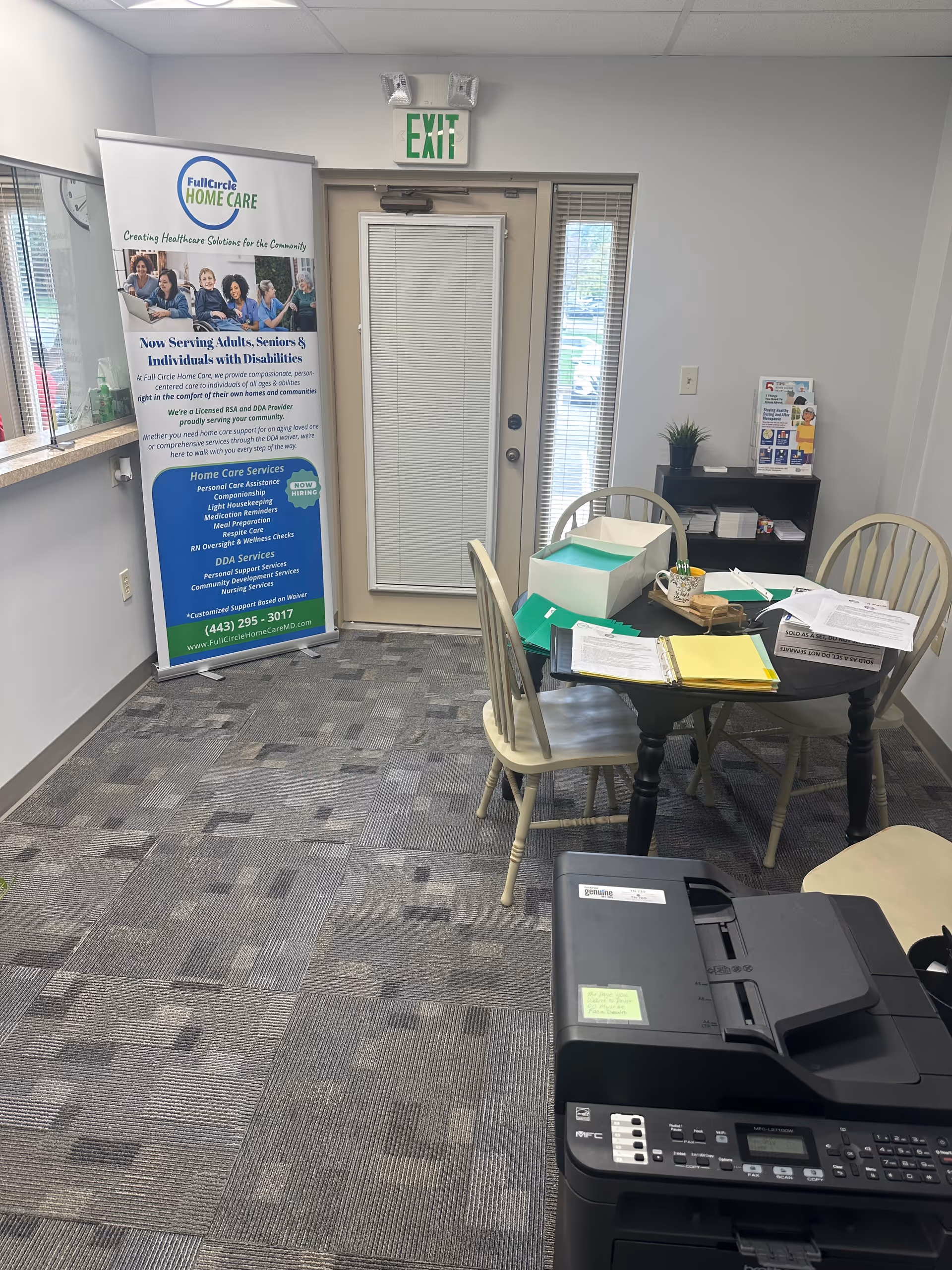 Indoor office space with a round table, chairs, a banner, exit sign, and a copier.