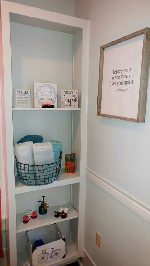 Tall white shelving unit with towels in a basket, decor signs, and framed quote.