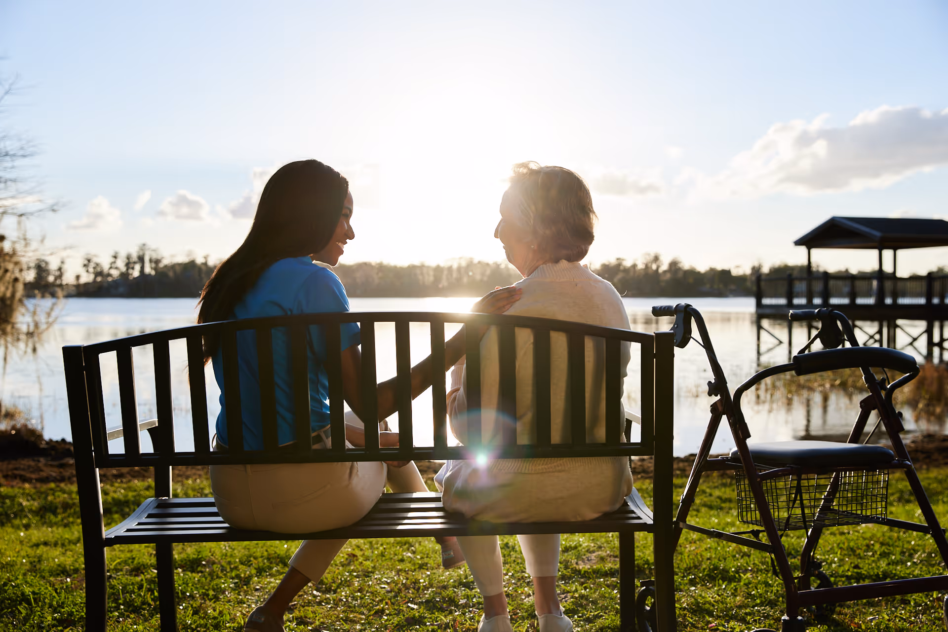 Two people sit on a bench beside a lake, sunlight glare, with a walker nearby.