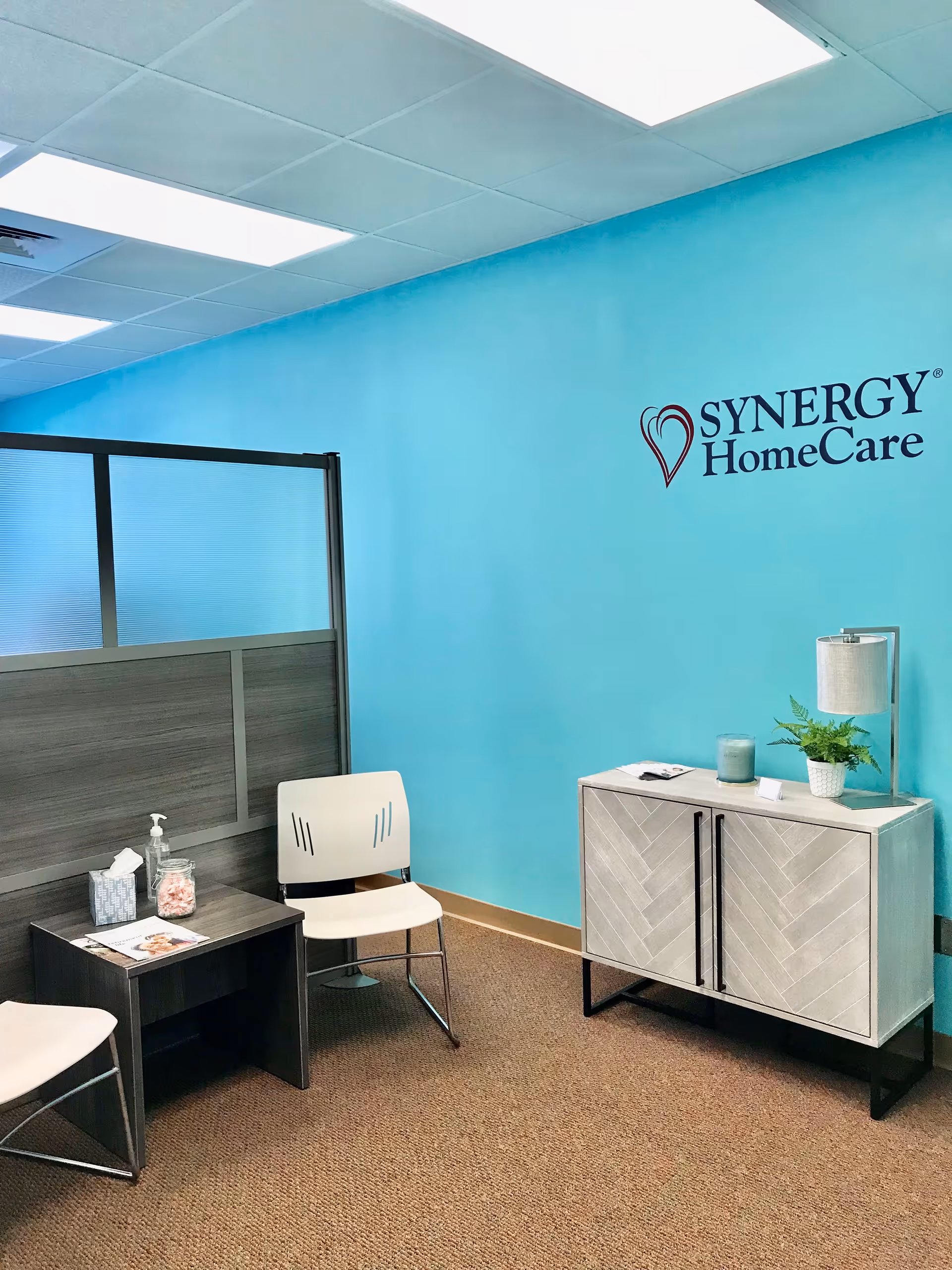 Bright turquoise reception area with chairs, a cabinet, and SYNERGY HomeCare logo.