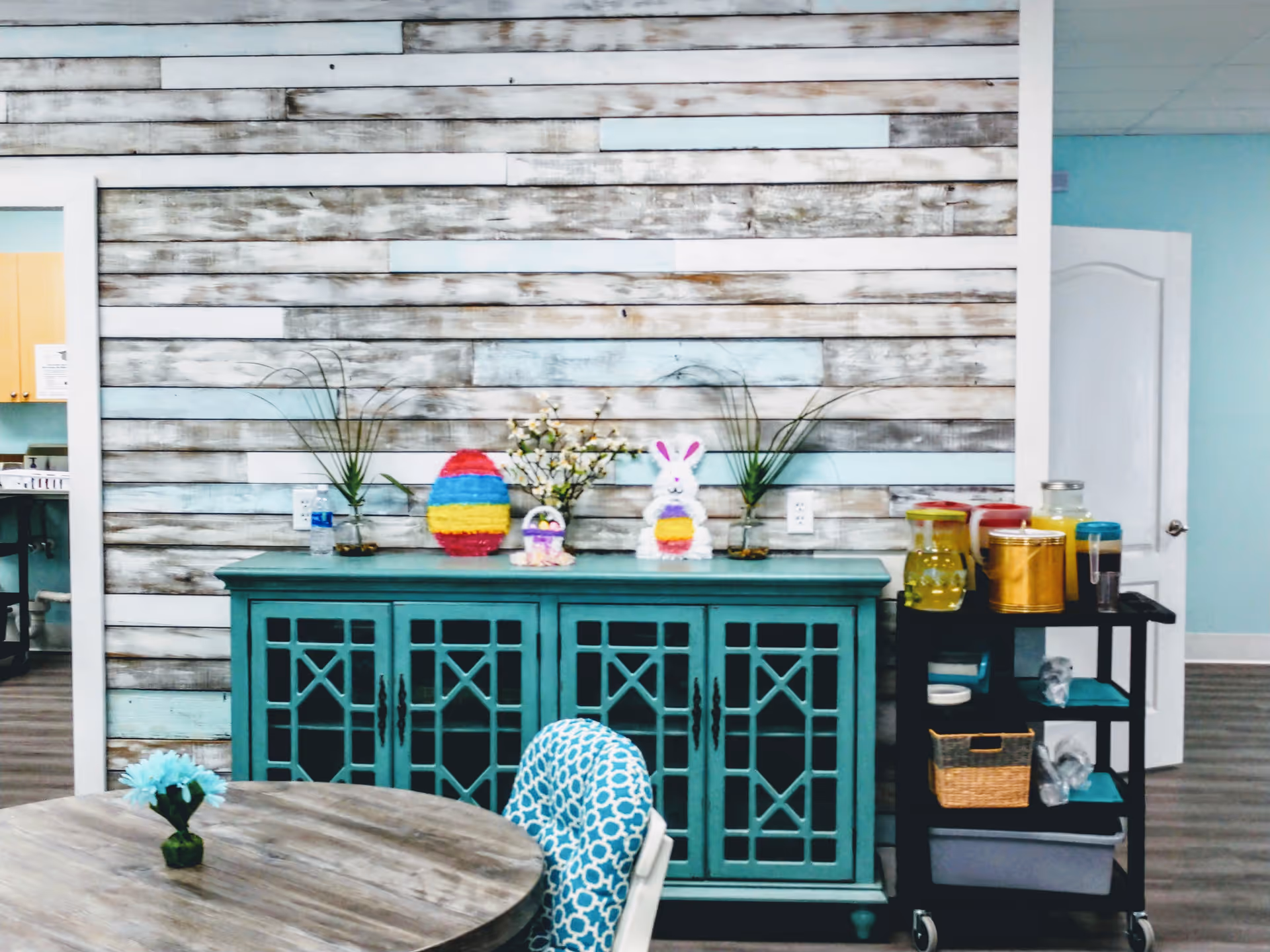 A teal cabinet against a rustic wood wall, with a round table and Easter decor.