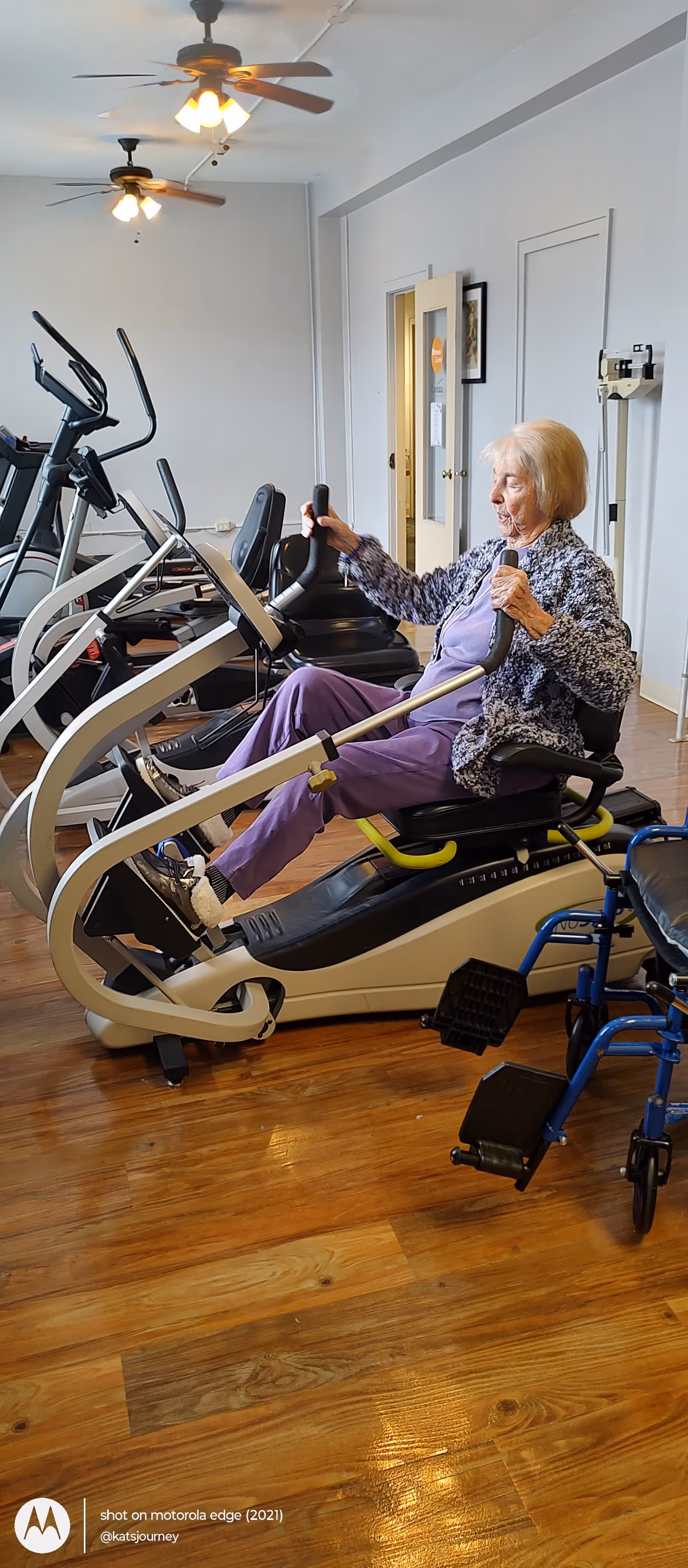 Elderly woman using a stationary bike in a bright indoor fitness room.