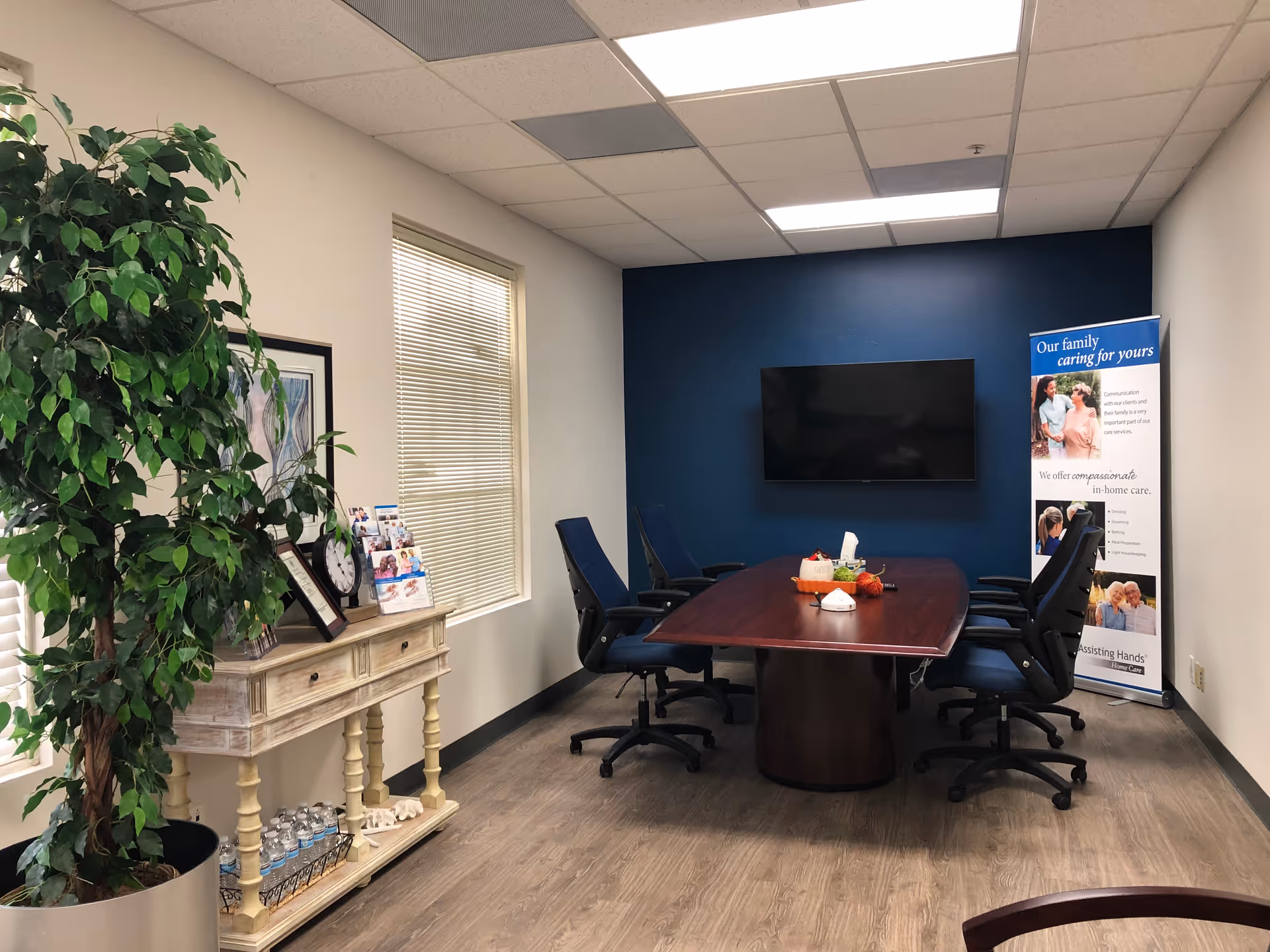 Conference room with a large table, blue wall, chairs, TV, banner, and plants.