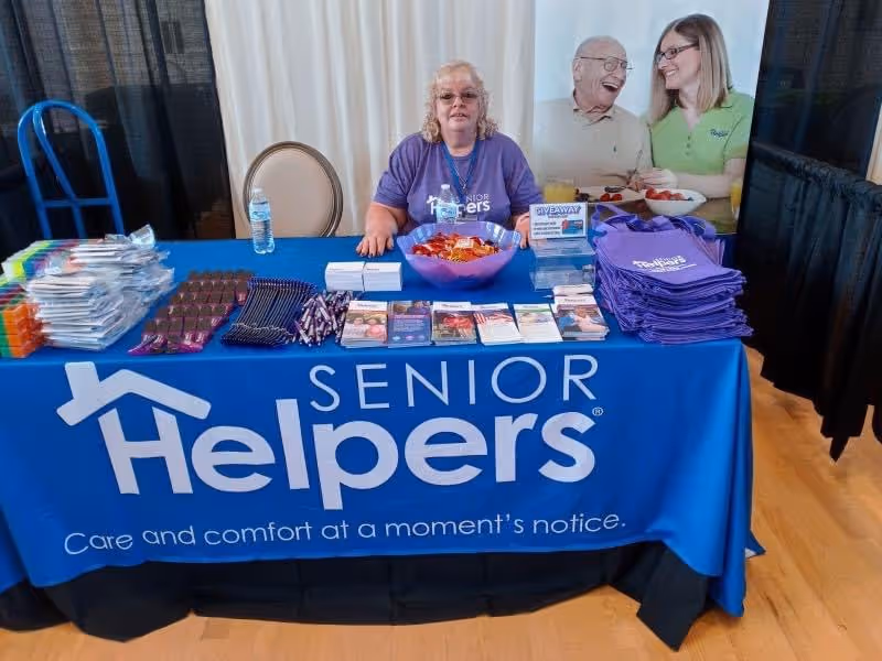 Indoors, a Senior Helpers promotional booth with staff behind a blue table covered in brochures.