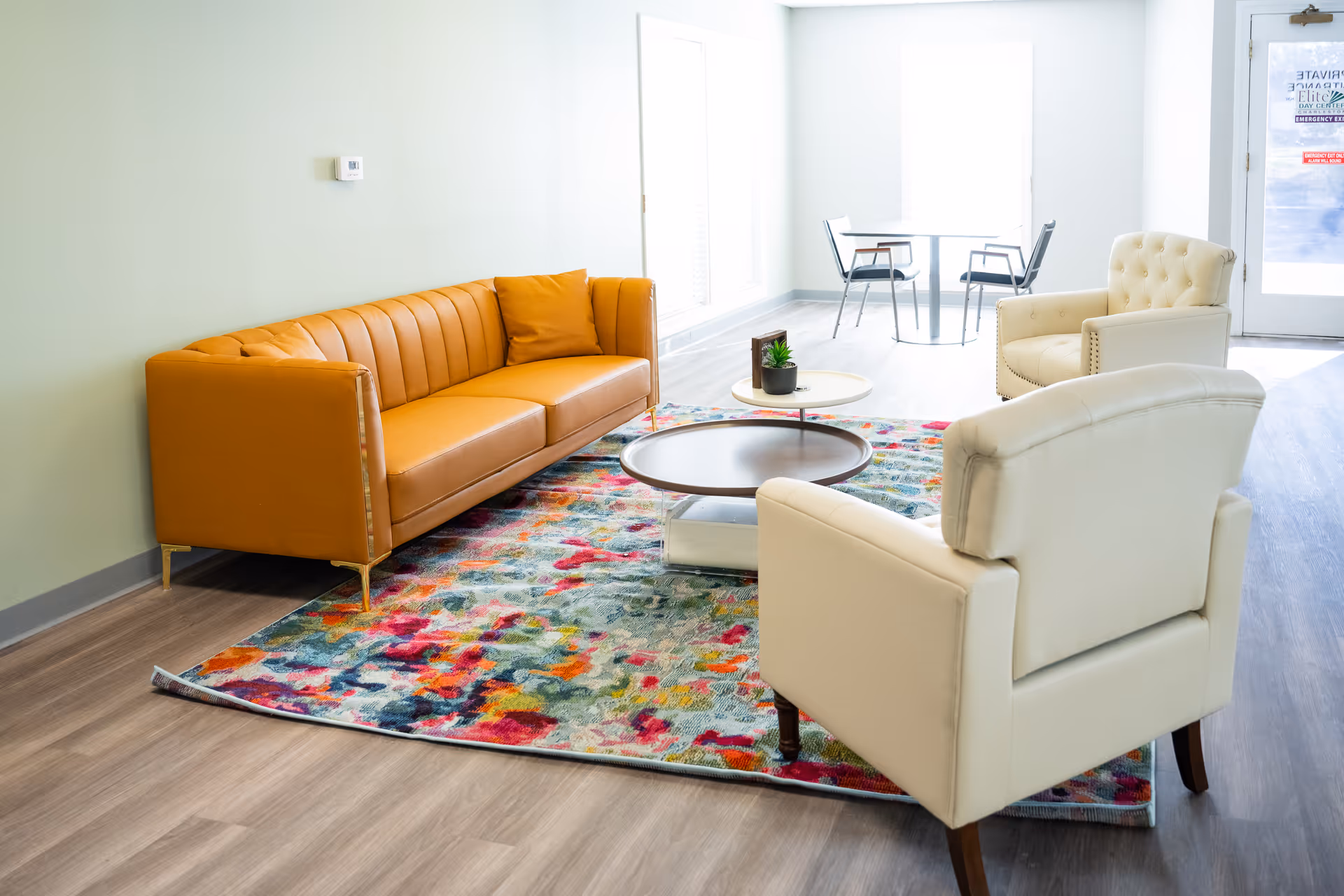 Sunlit senior living lounge with orange sofa, cream chairs, colorful rug, and coffee table.