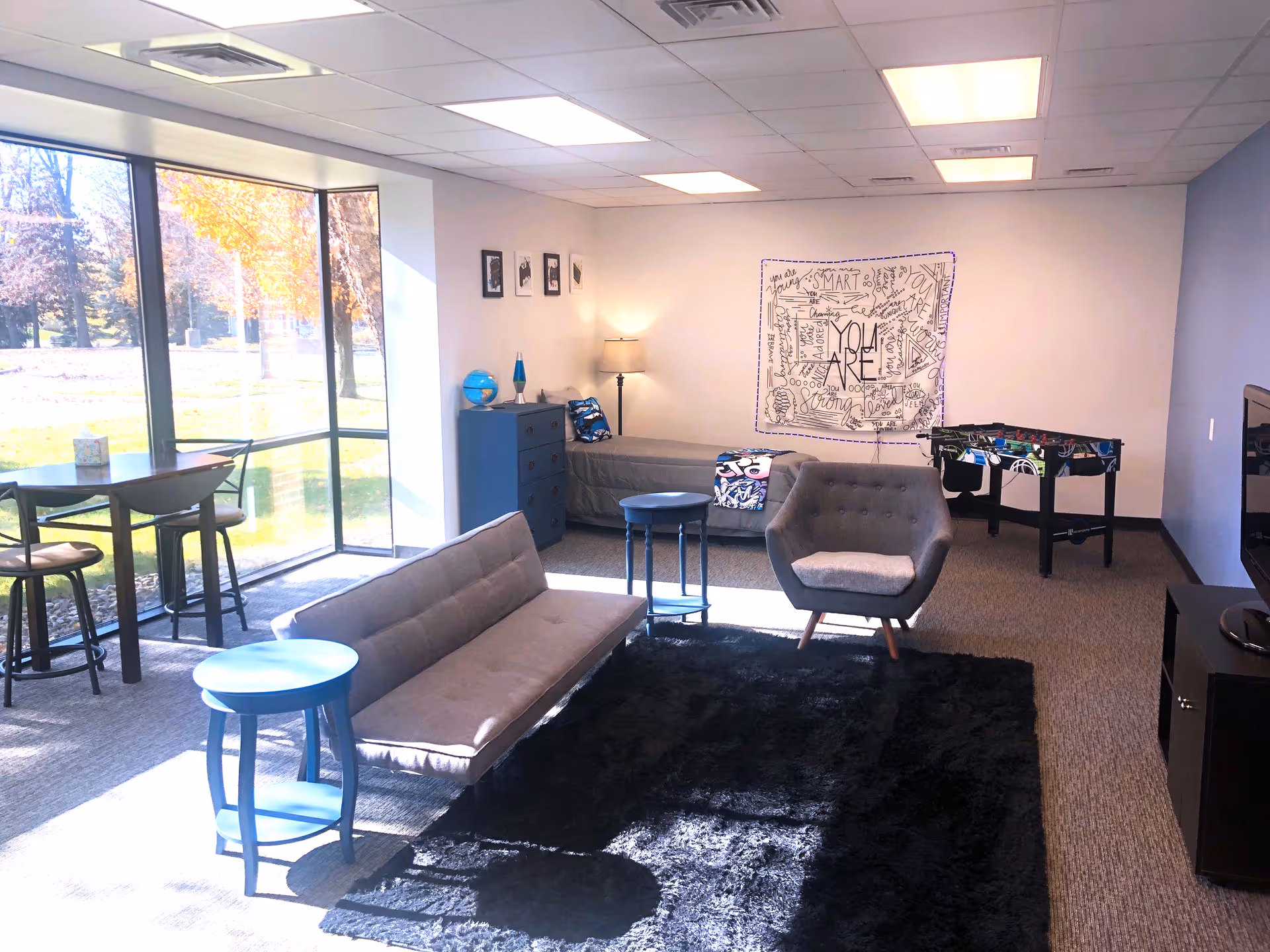 Bright lounge area with seating, tables, and a foosball table by large windows.