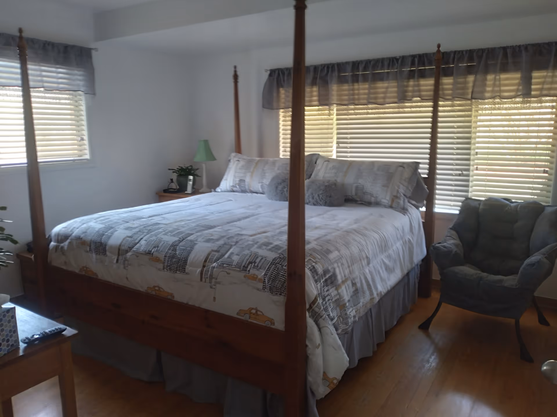 A four-poster bed dominates a cozy bedroom with blinds, nightstand, lamp, and chair.