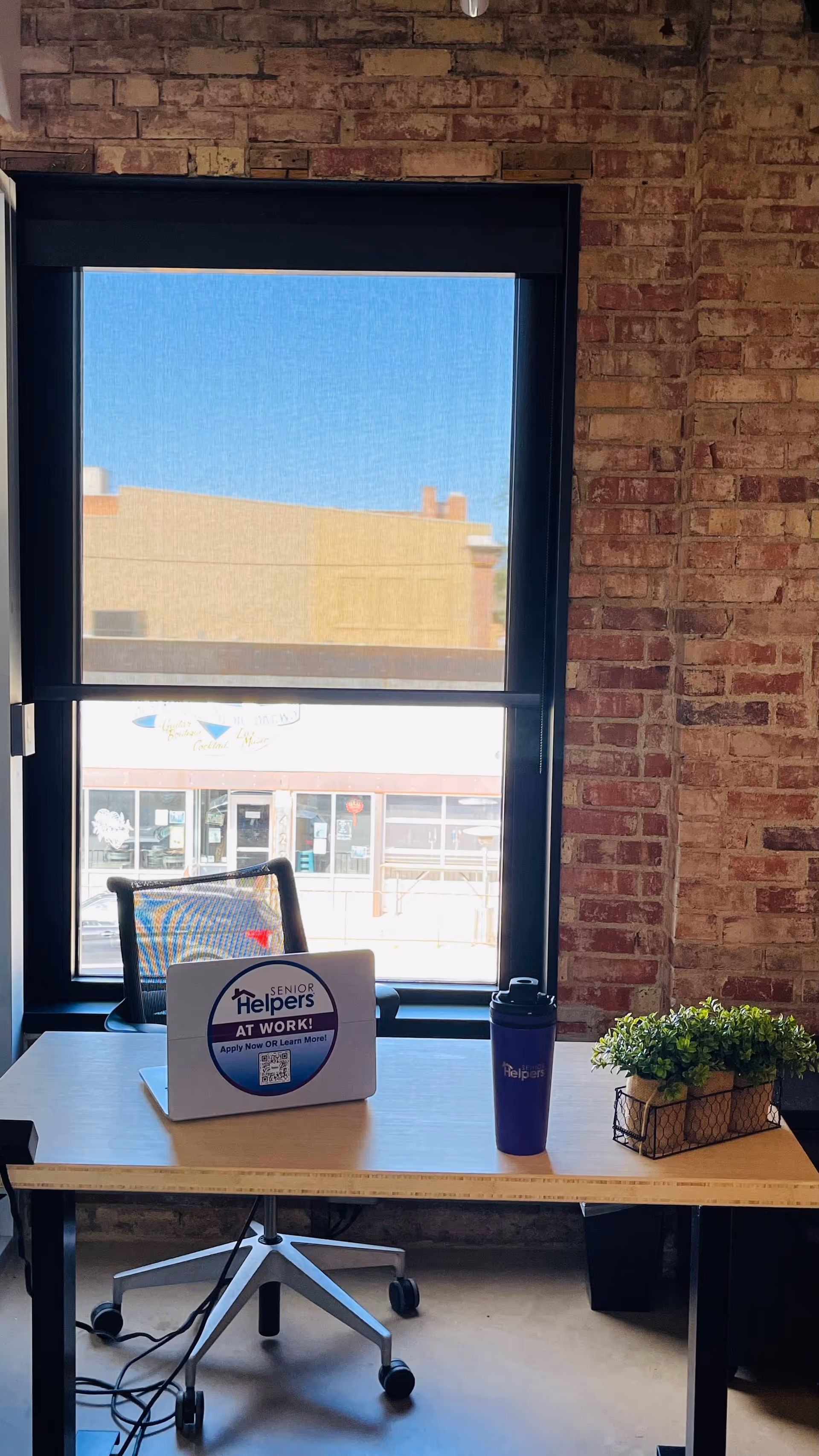 Office desk by a large window with brick wall, chair, sign, mug, and plant.