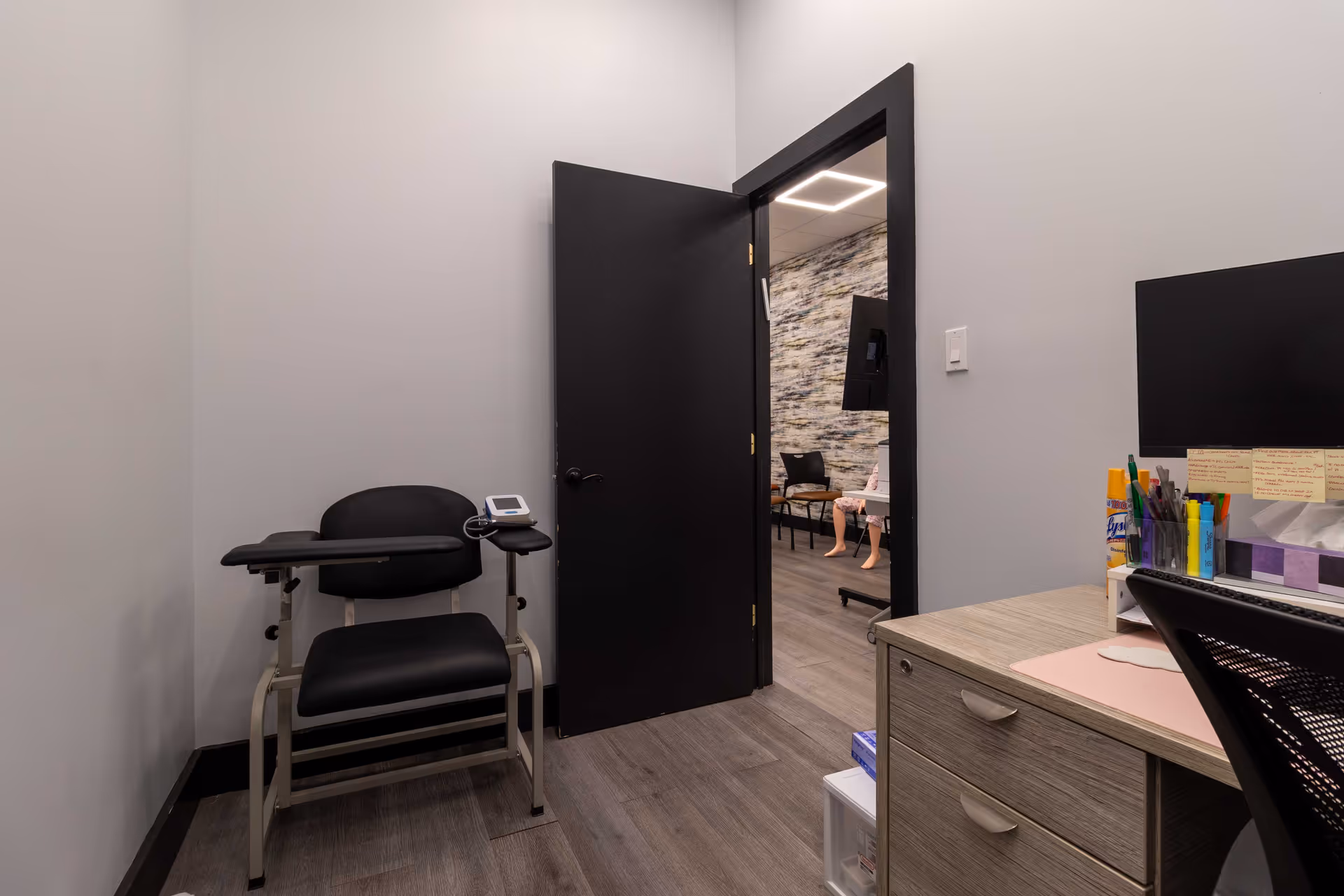 Empty medical examination room with a chair, desk, and doorway to another area.