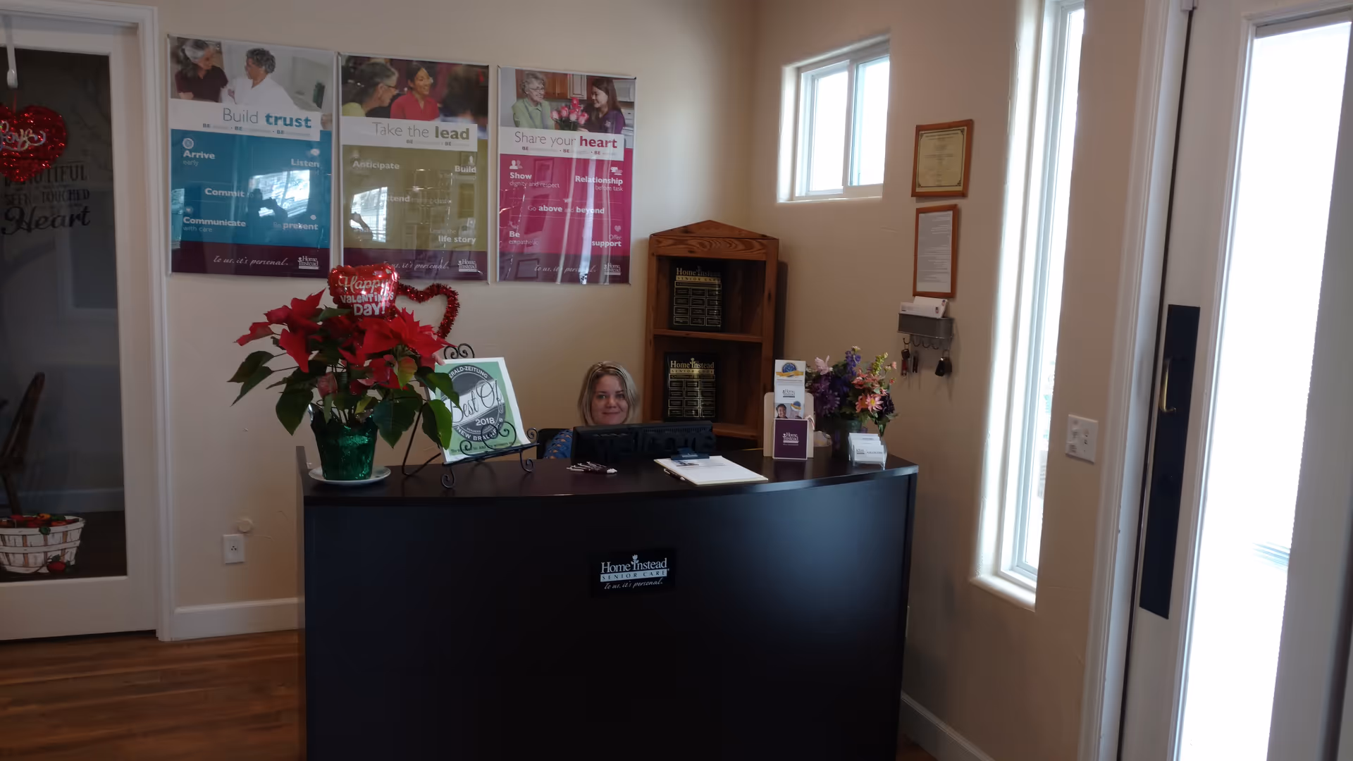 Reception desk with a smiling staff member in a lobby, posters, flowers, and windows.