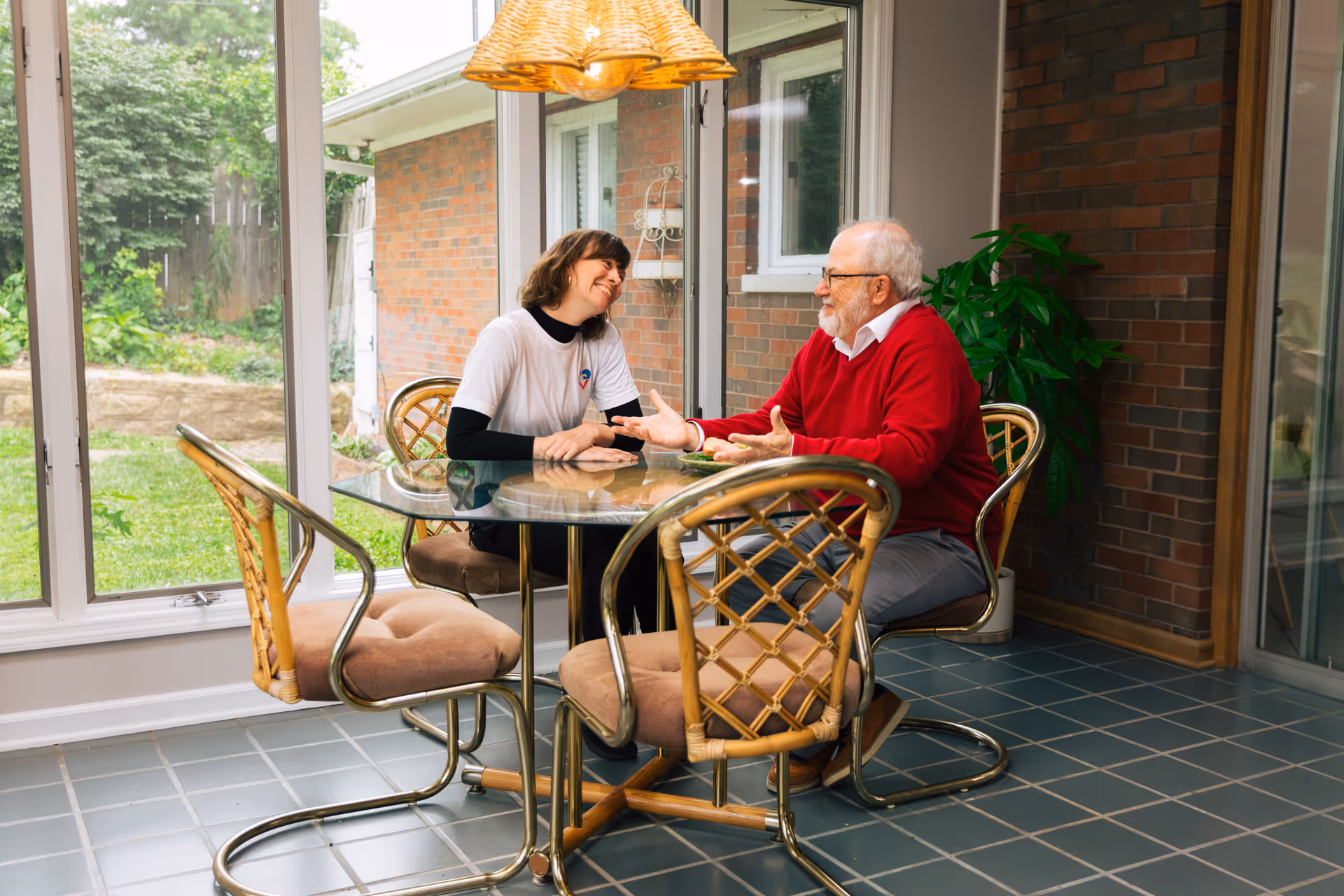 Two people converse at a glass table in a bright sunlit living area.