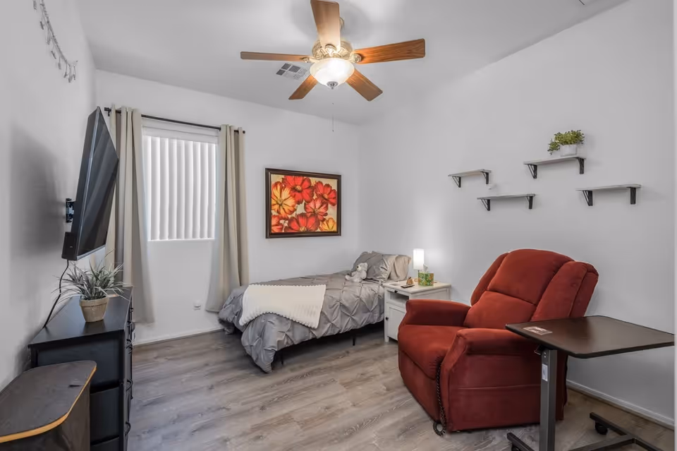 A small, tidy bedroom with a bed, red recliner, TV, nightstand, and wall shelves.