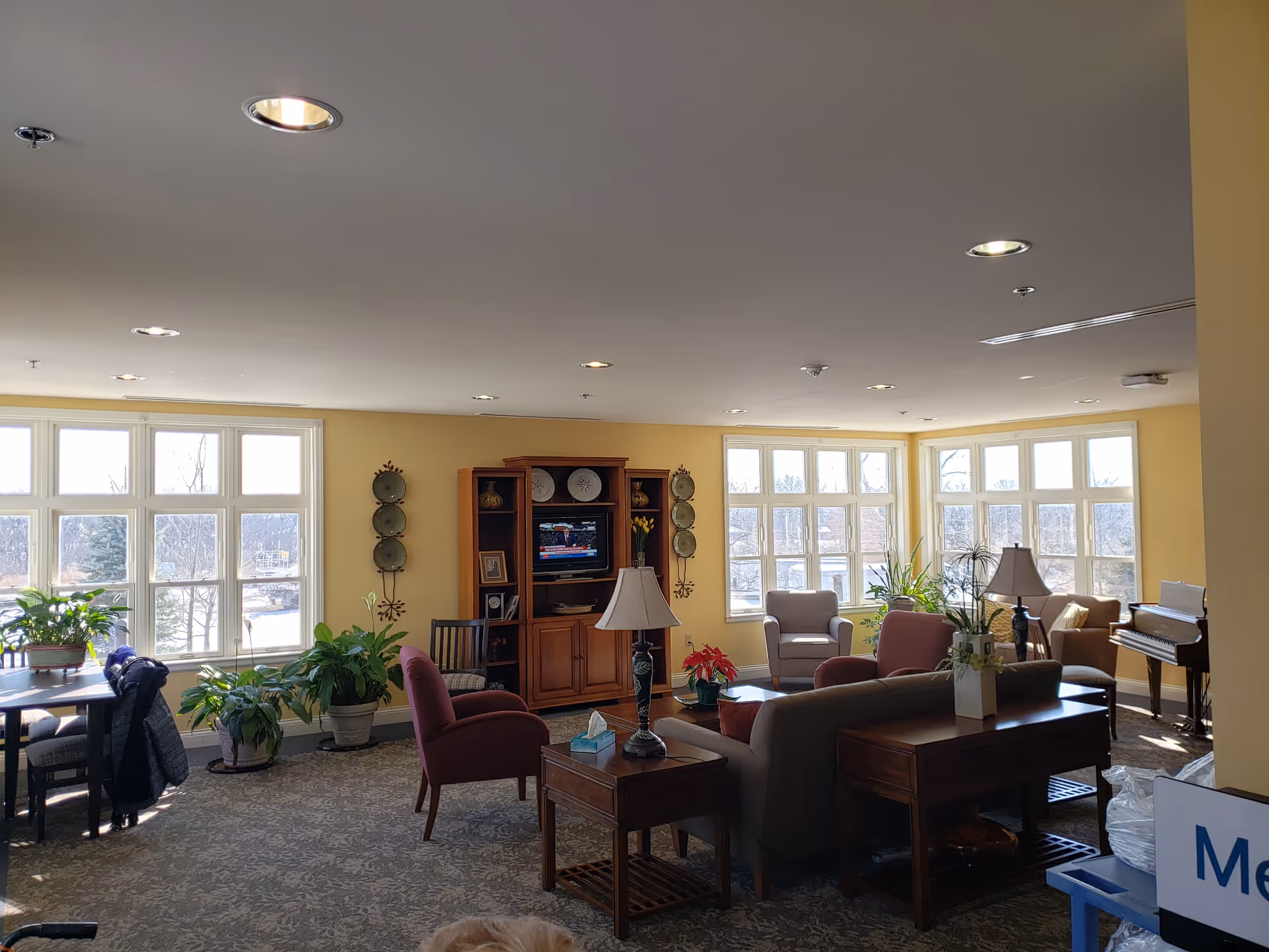 Bright communal living room with multiple seating areas, plants, a TV cabinet, and a piano.
