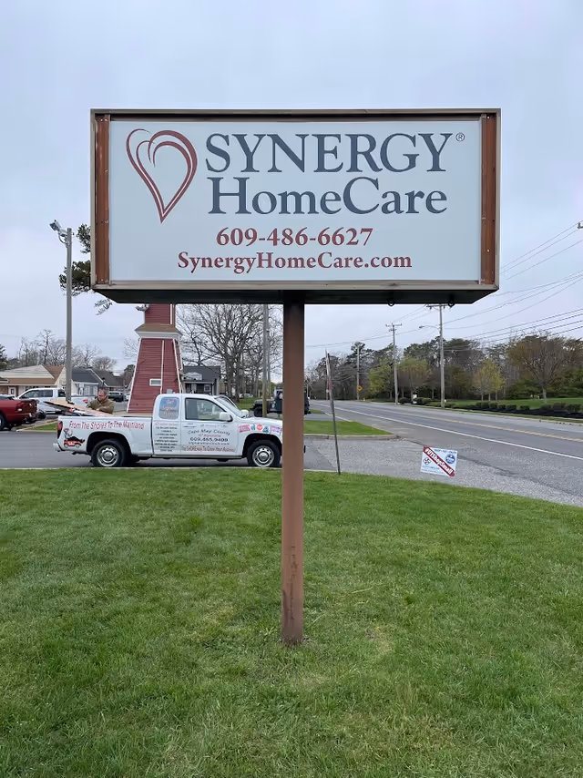 Large SYNERGY HomeCare sign on a wooden post near a grassy lawn and road.