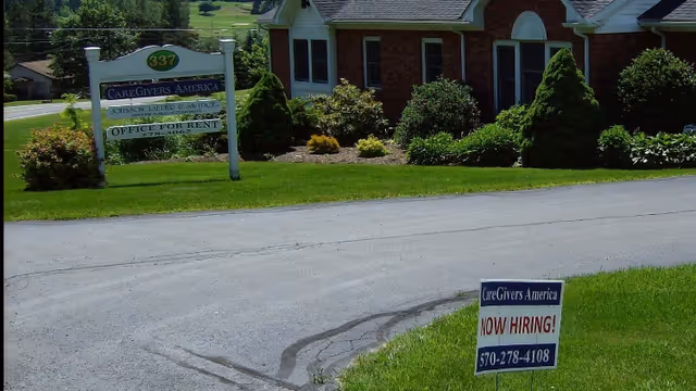Exterior view of CareGivers America facility with lawn signs and Now Hiring poster