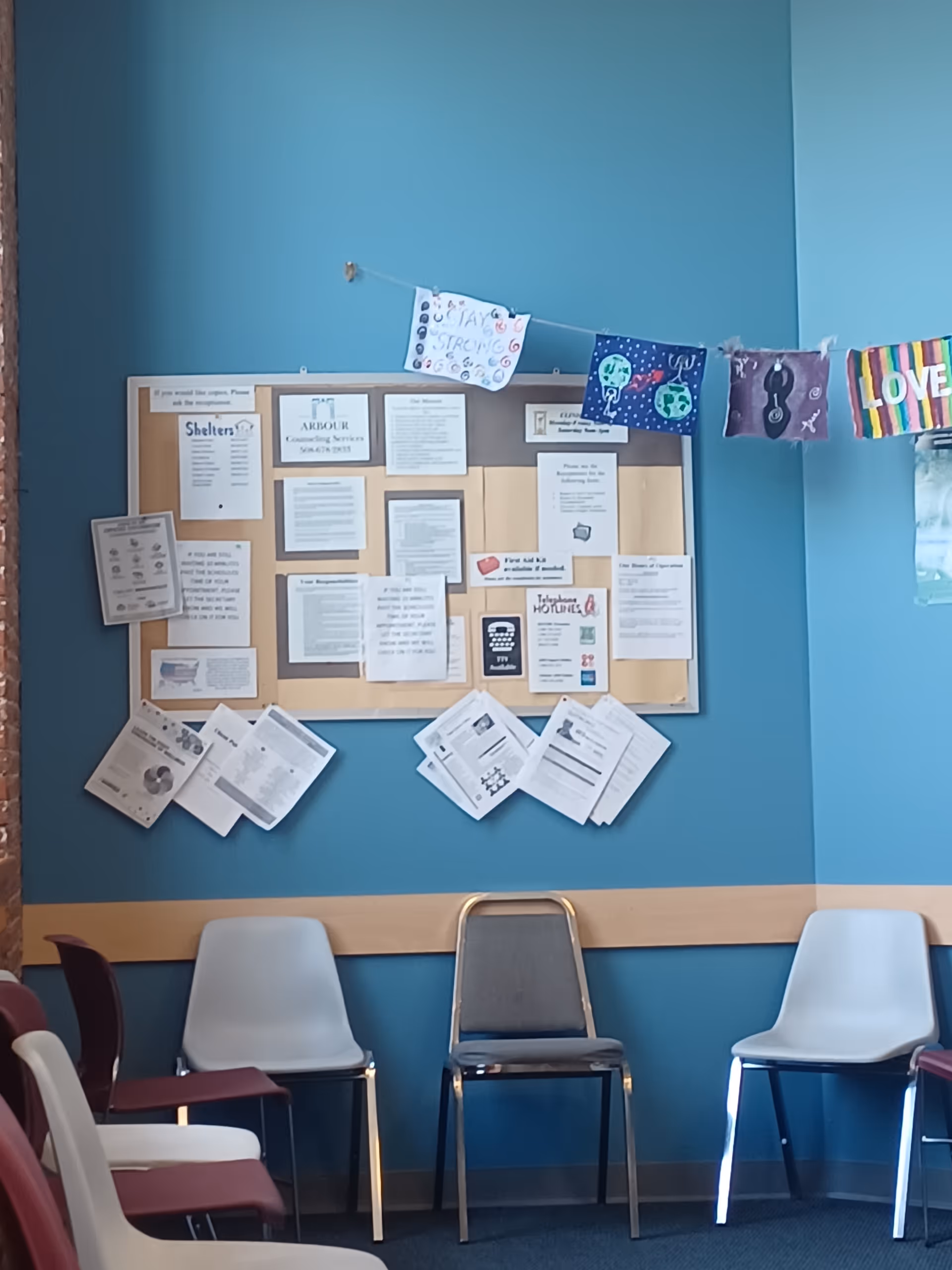 Blue-walled waiting area with bulletin board of notices and several plastic chairs.