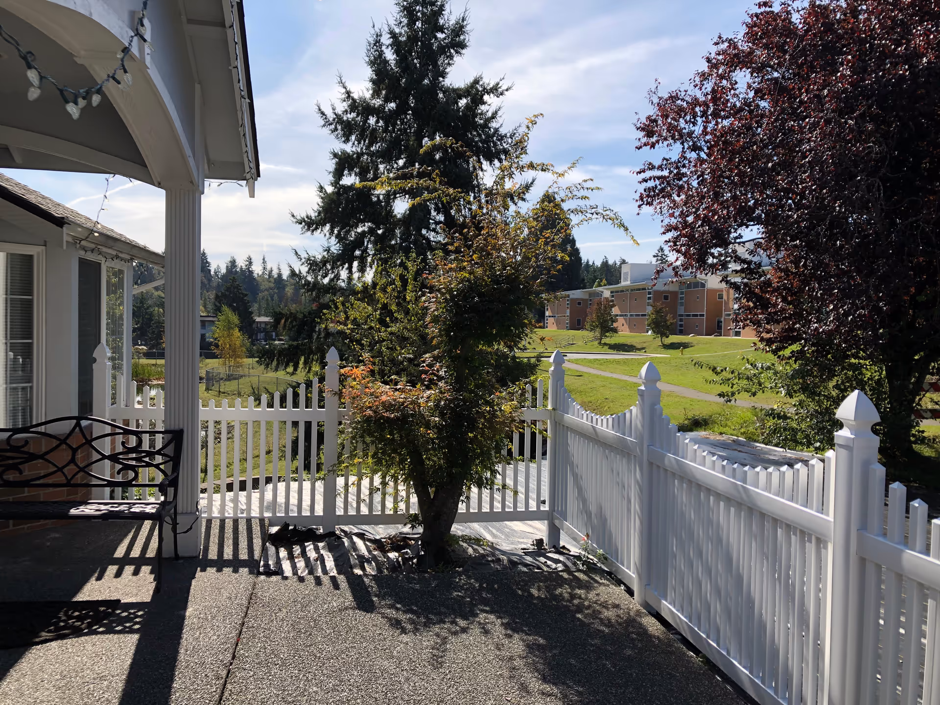 Sunlit porch with a white fence, bench, and trees overlooking a distant senior living building.